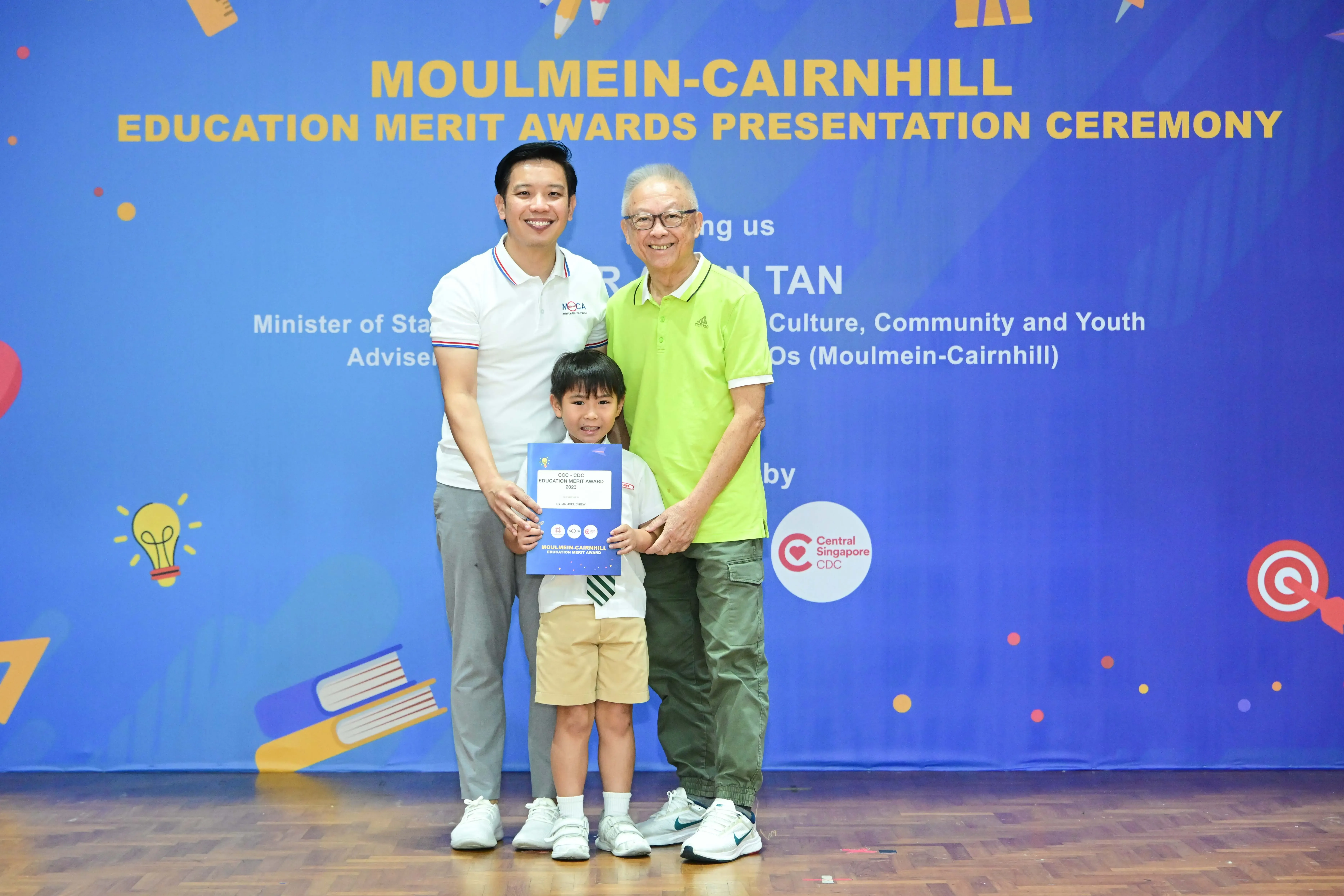 Two men and a young child holding a certificate pose before a Moulmein-Cairnhill Education Merit Awards banner.