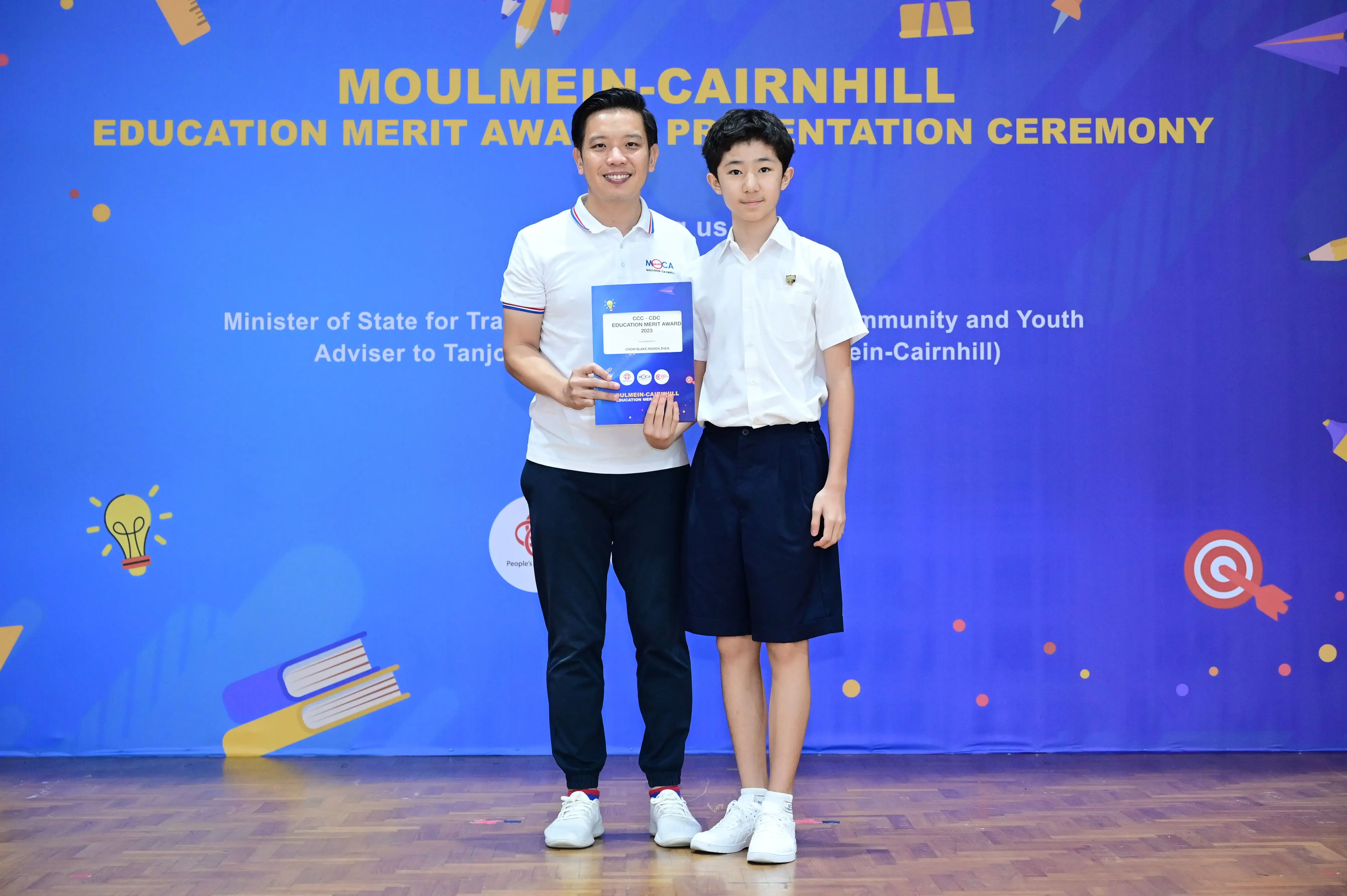An adult and a young student pose together holding an award certificate at the Moulmein-Cairnhill Education Merit Award ceremony.