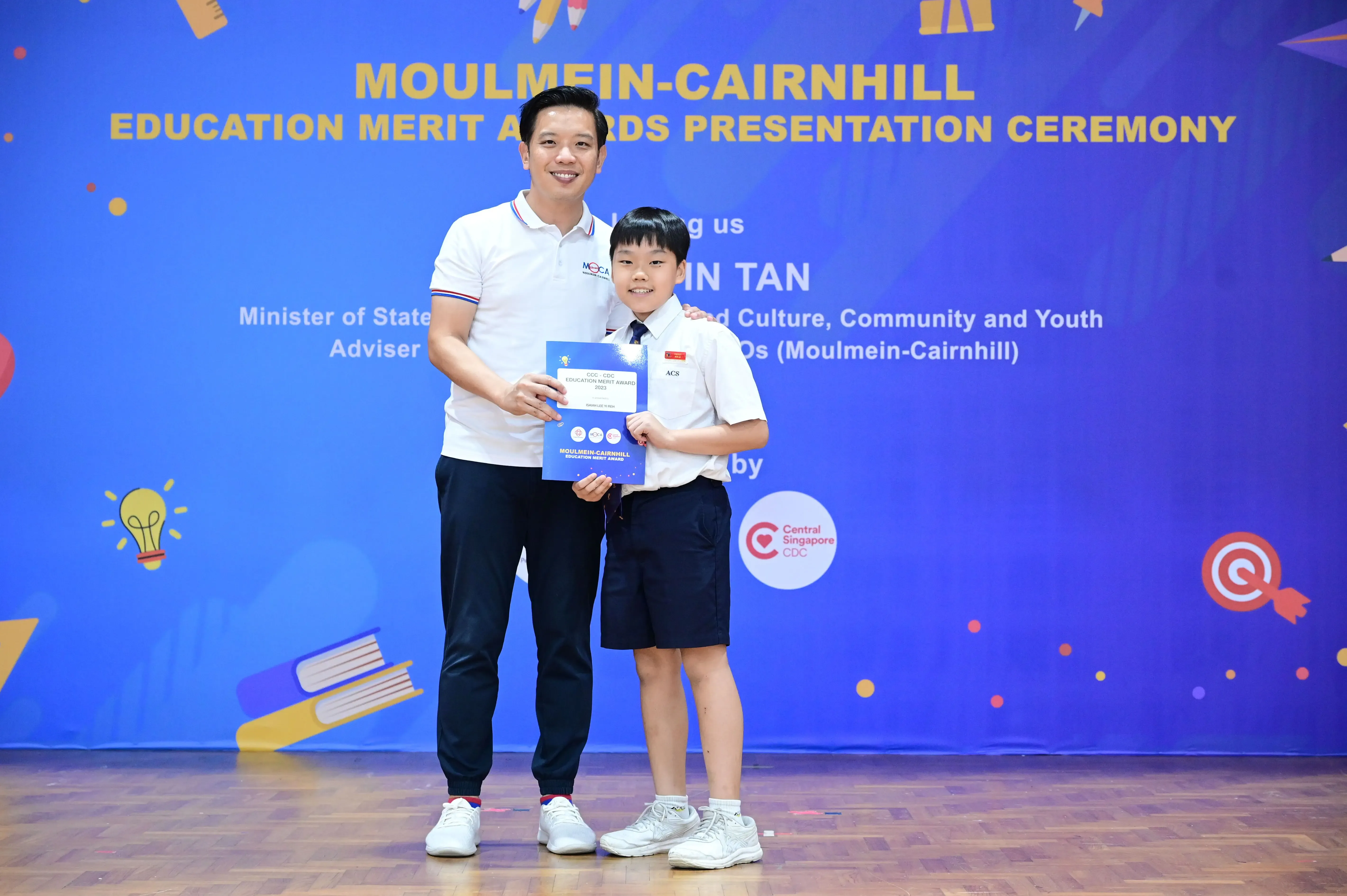 A man and a young student in uniform hold a certificate at the Moulmein-Cairnhill Education Merit Awards ceremony.