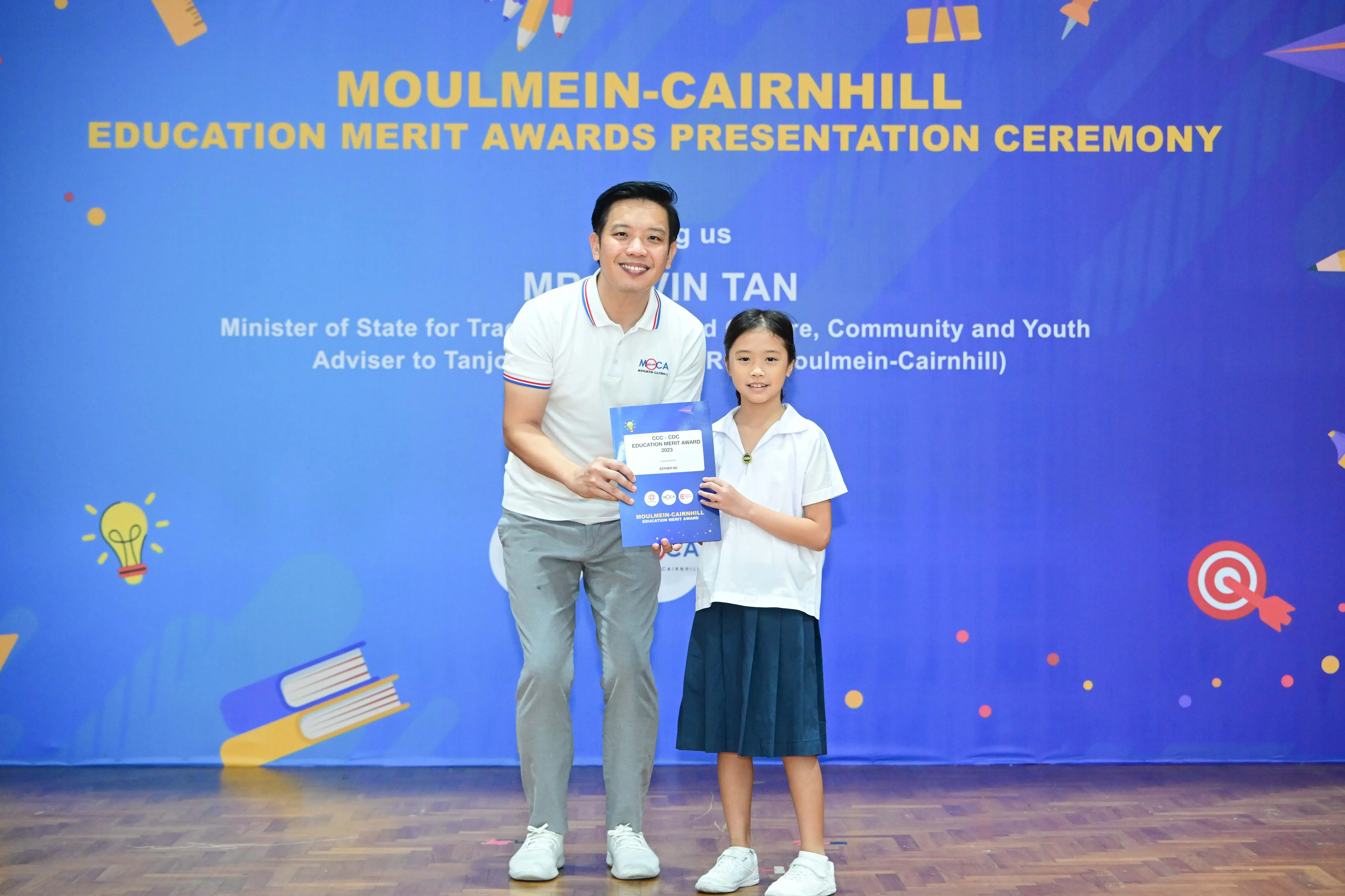 A man and a young girl hold a blue award booklet at the Moulmein-Cairnhill Education Merit Awards Ceremony.