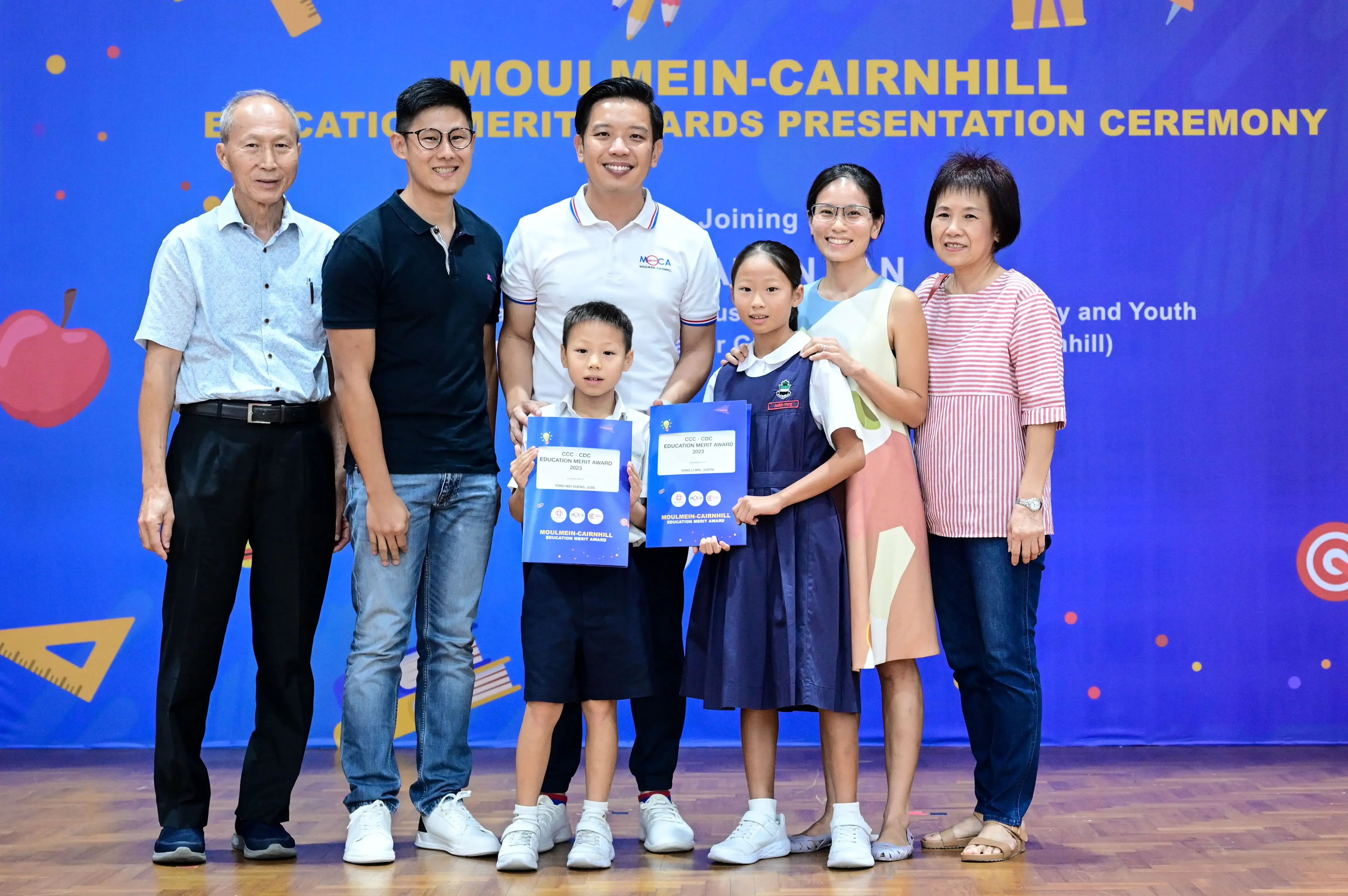 Seven people pose at the Moulmein-Cairnhill Education Merit Awards Presentation Ceremony, two children holding blue award certificates.