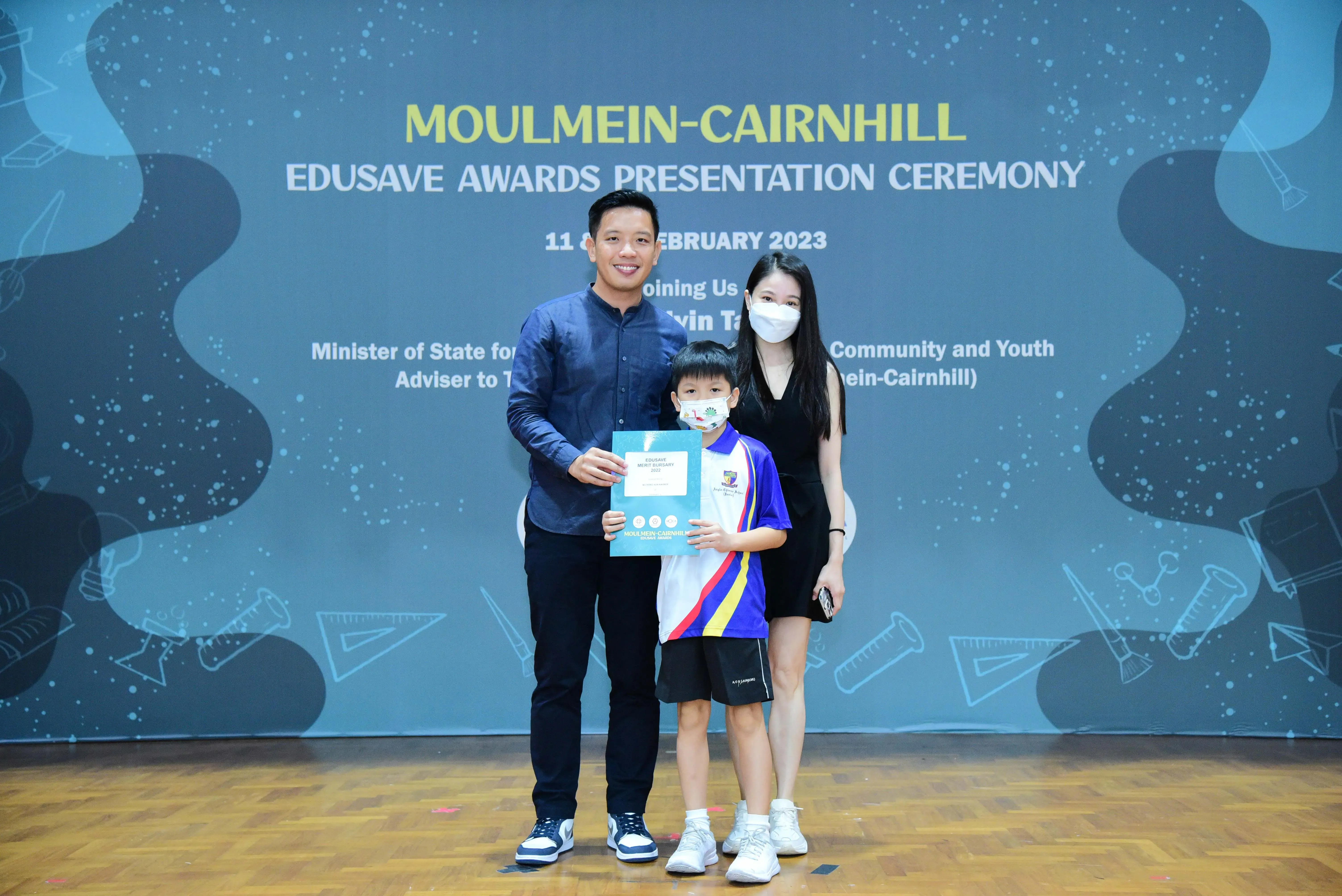 Two adults and a young boy holding a certificate pose at the Moulmein-Cairnhill Edusave Awards Ceremony 2023.
