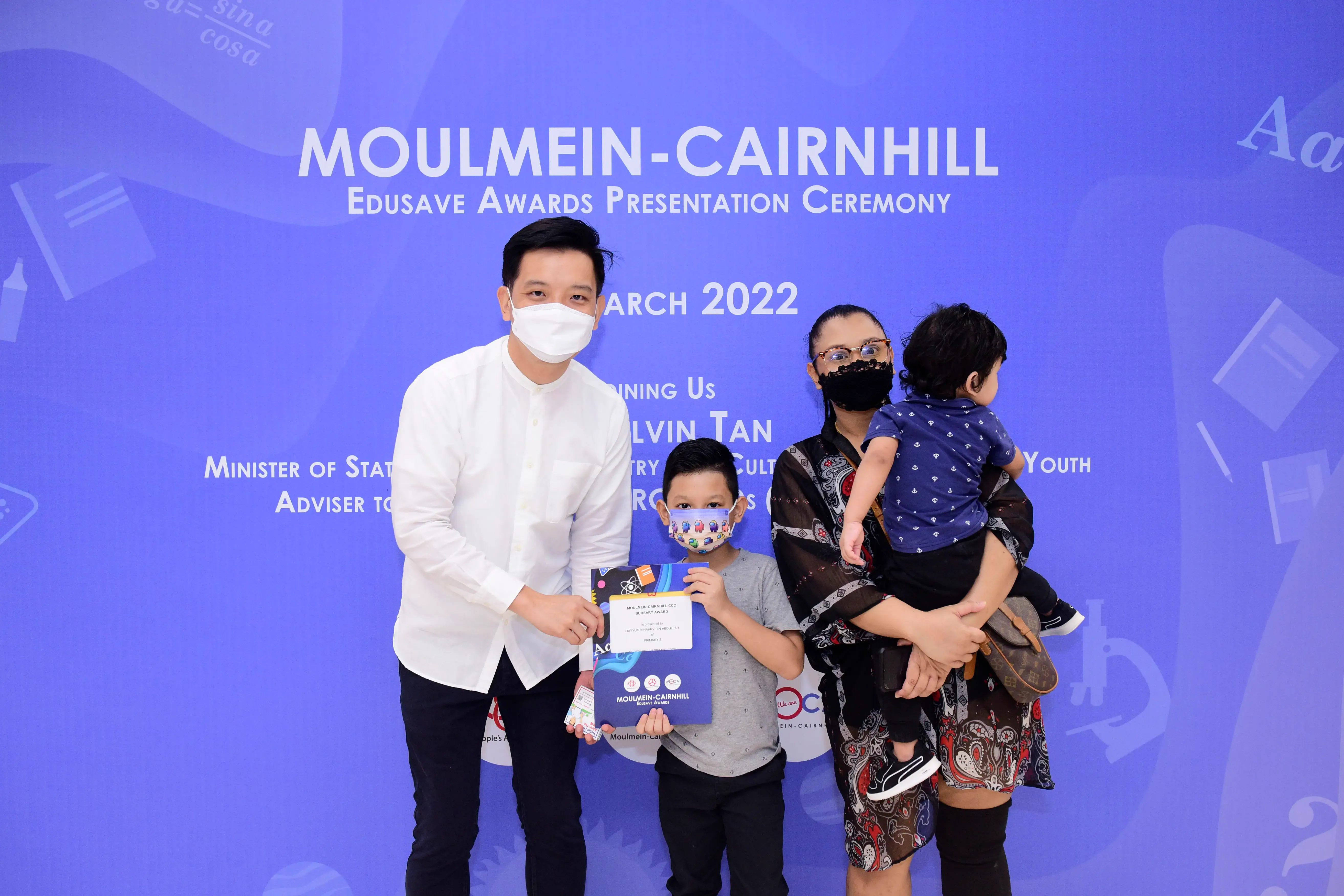 Family of four wearing masks posing at the Moulmein-Cairnhill Edusave Awards Ceremony 2022, child holding certificate.
