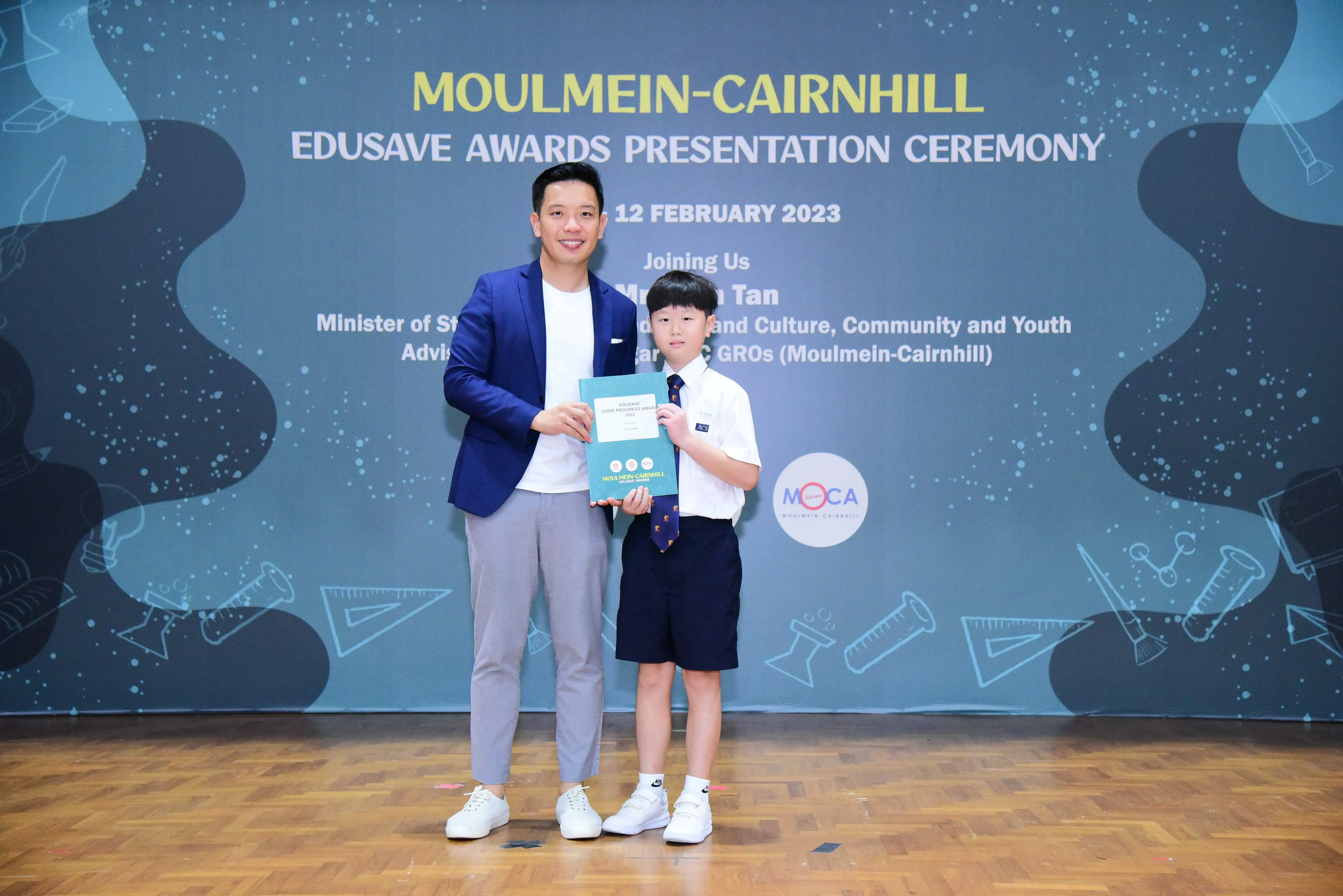 An adult in a blue blazer and a young student in uniform hold an award at the Moulmein-Cairnhill Edusave Awards Ceremony 2023.