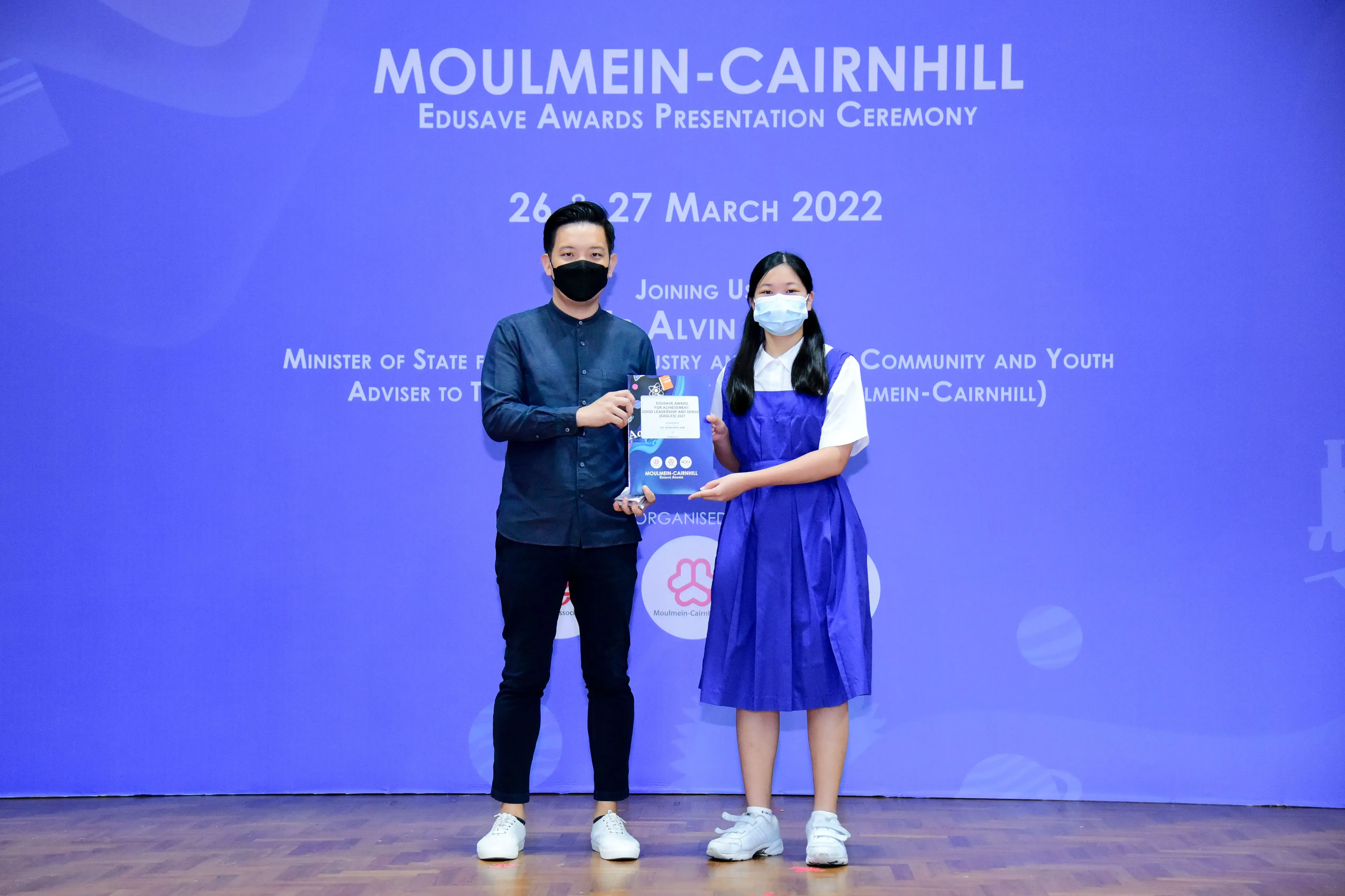 Two masked individuals hold a certificate at the Moulmein-Cairnhill Edusave Awards Presentation Ceremony, March 2022.