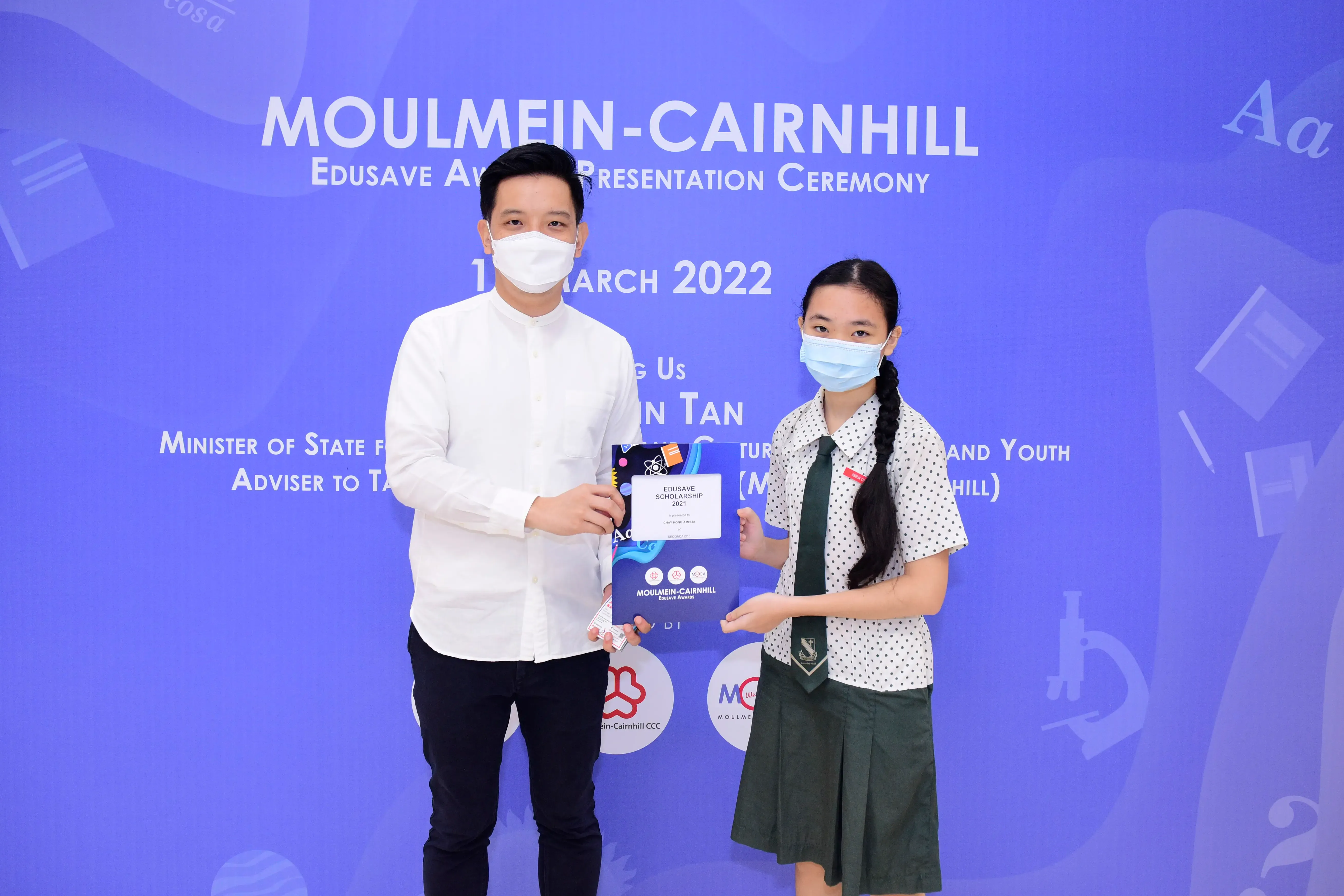 Two masked people hold an Edusave Scholarship certificate at the Moulmein-Cairnhill Awards Ceremony, March 2022.