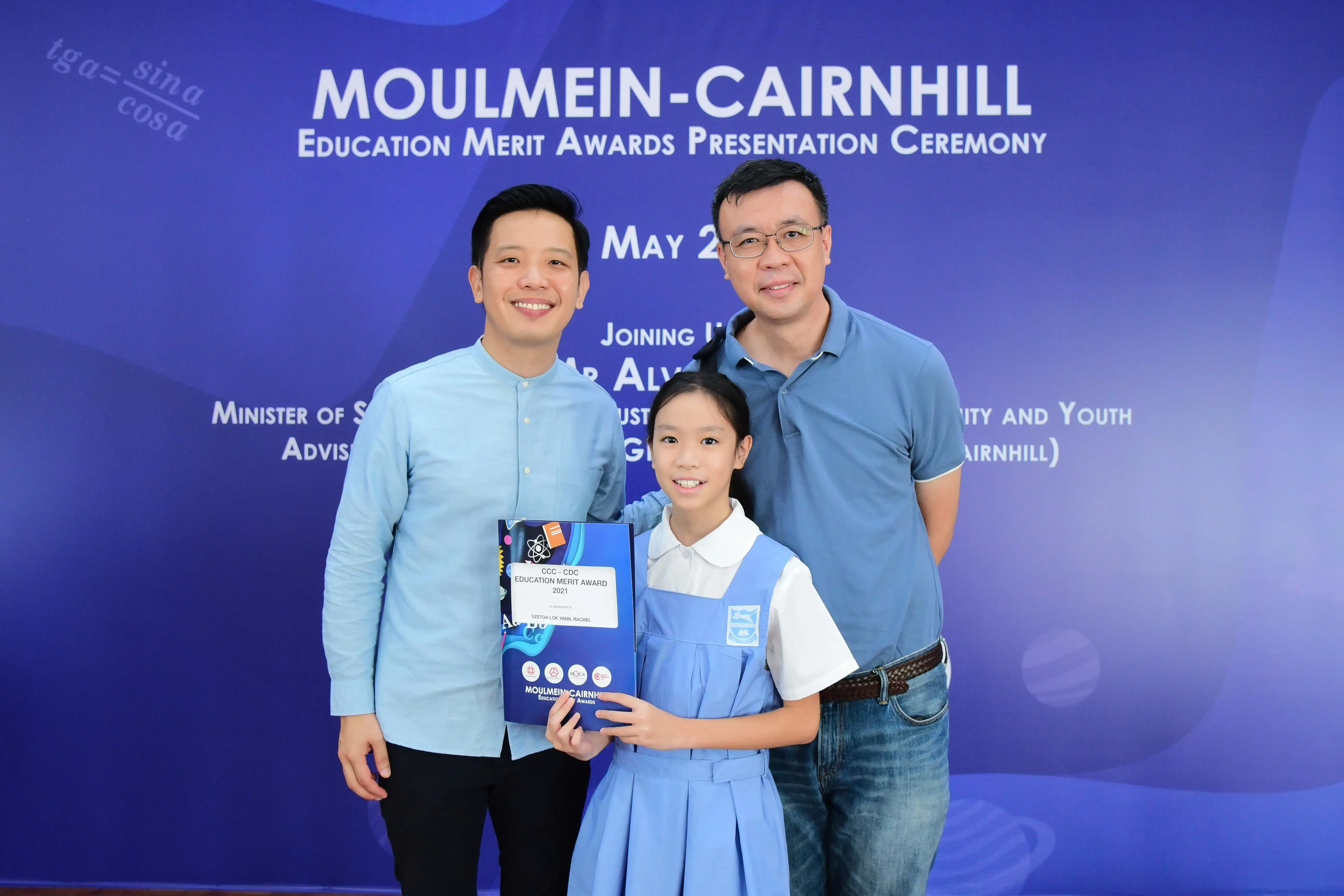 Two men and a girl in school uniform hold an award at the Moulmein-Cairnhill Education Merit Awards Ceremony.