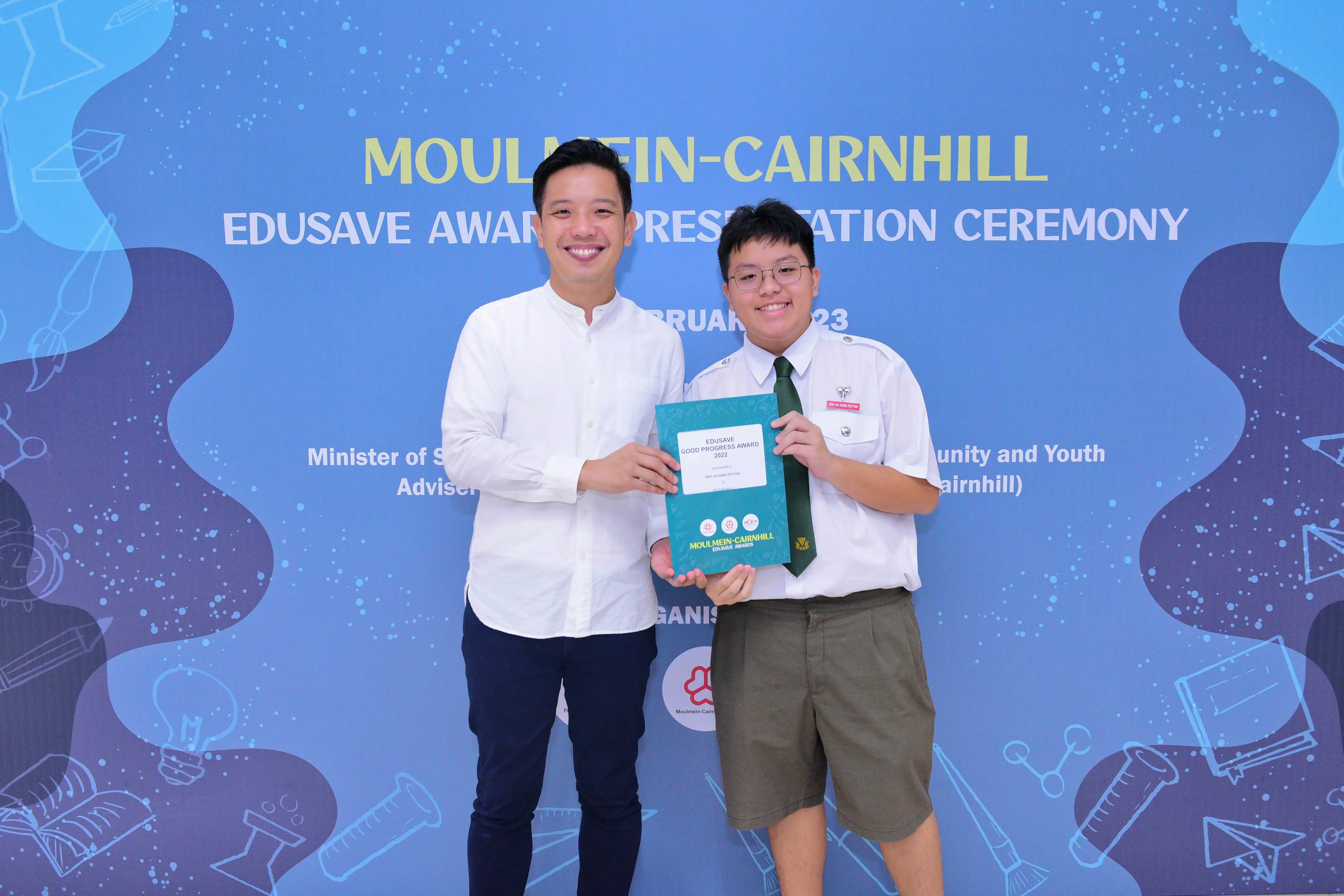 Two smiling people hold a teal award booklet at the Moulmein-Cairnhill Edusave Award Presentation Ceremony.