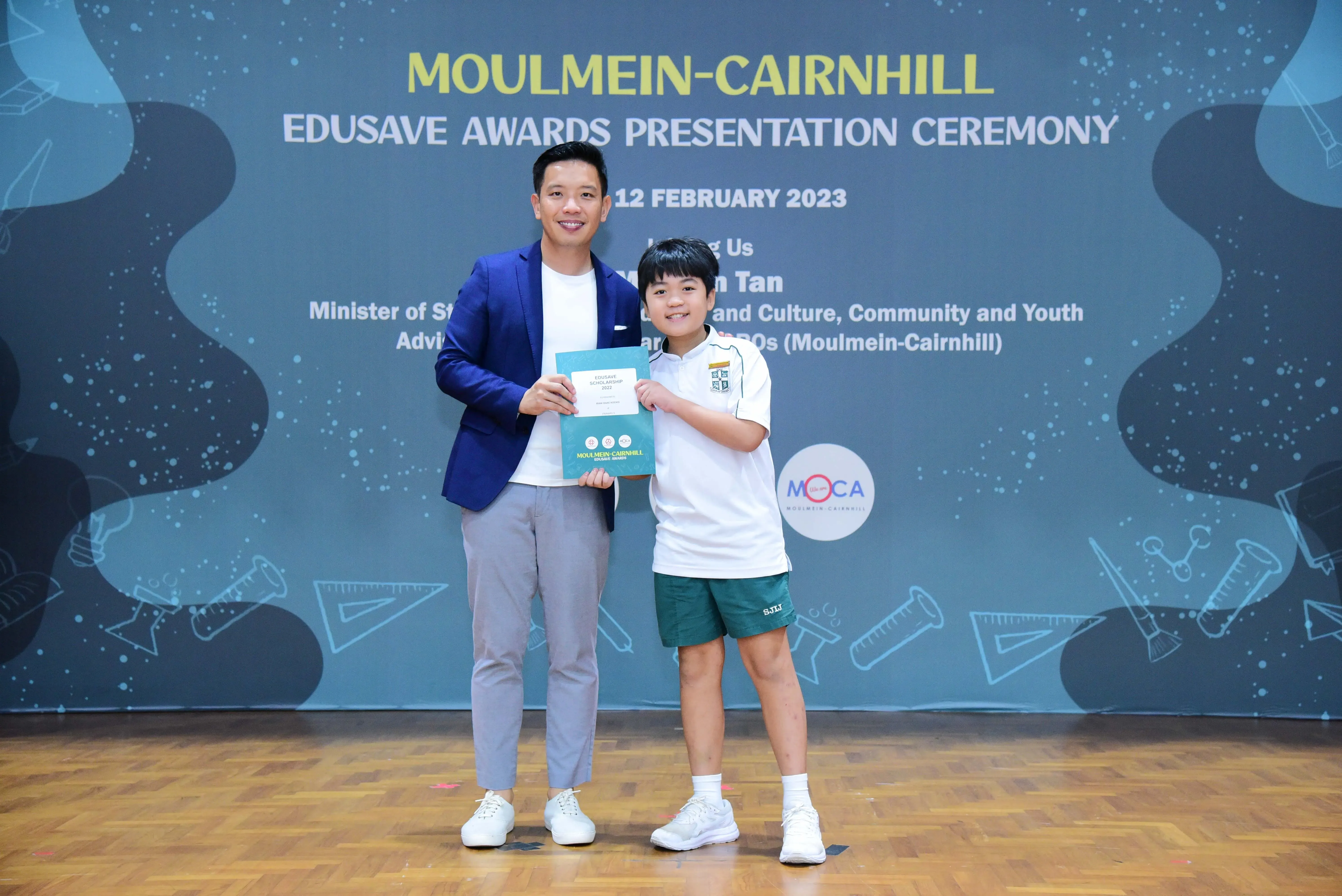 An adult in a blue blazer and a young student hold a certificate at the Moulmein-Cairnhill Edusave Awards Ceremony 2023.