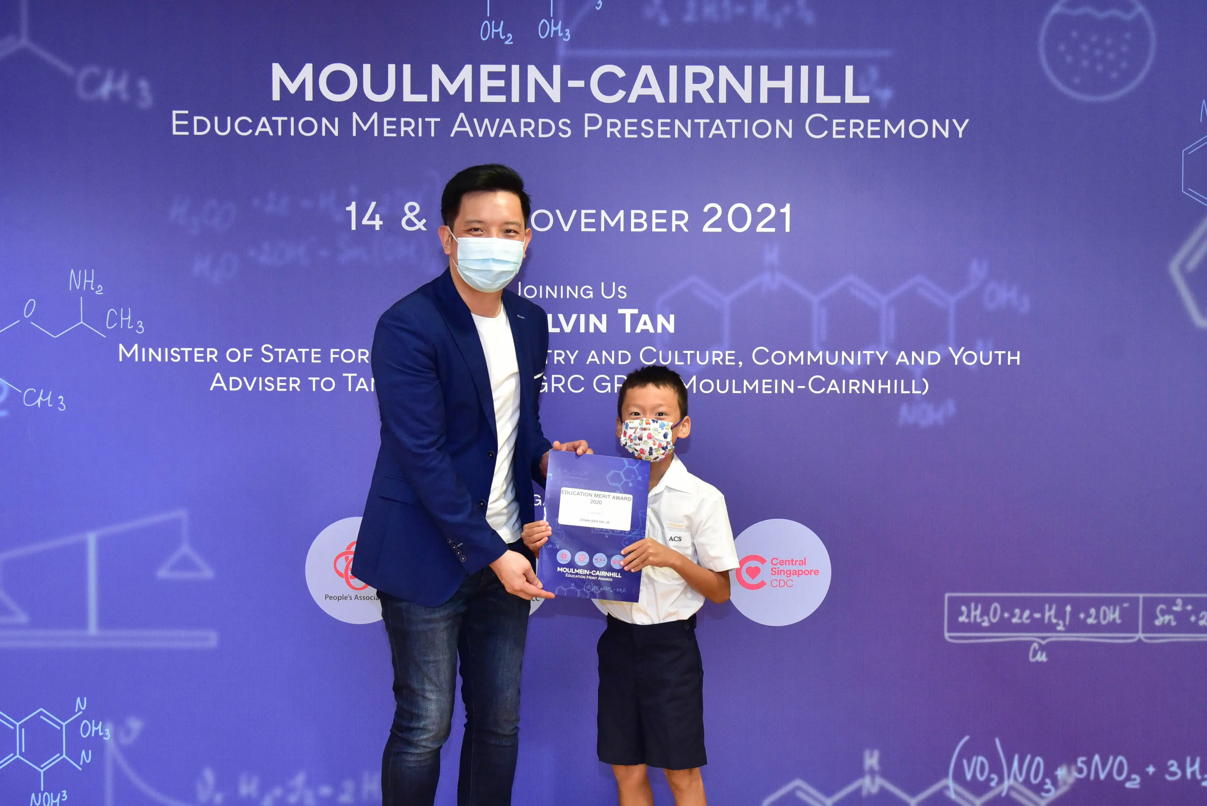 An adult and a young boy in masks hold an award certificate at the Moulmein-Cairnhill Education Merit Awards 2021.