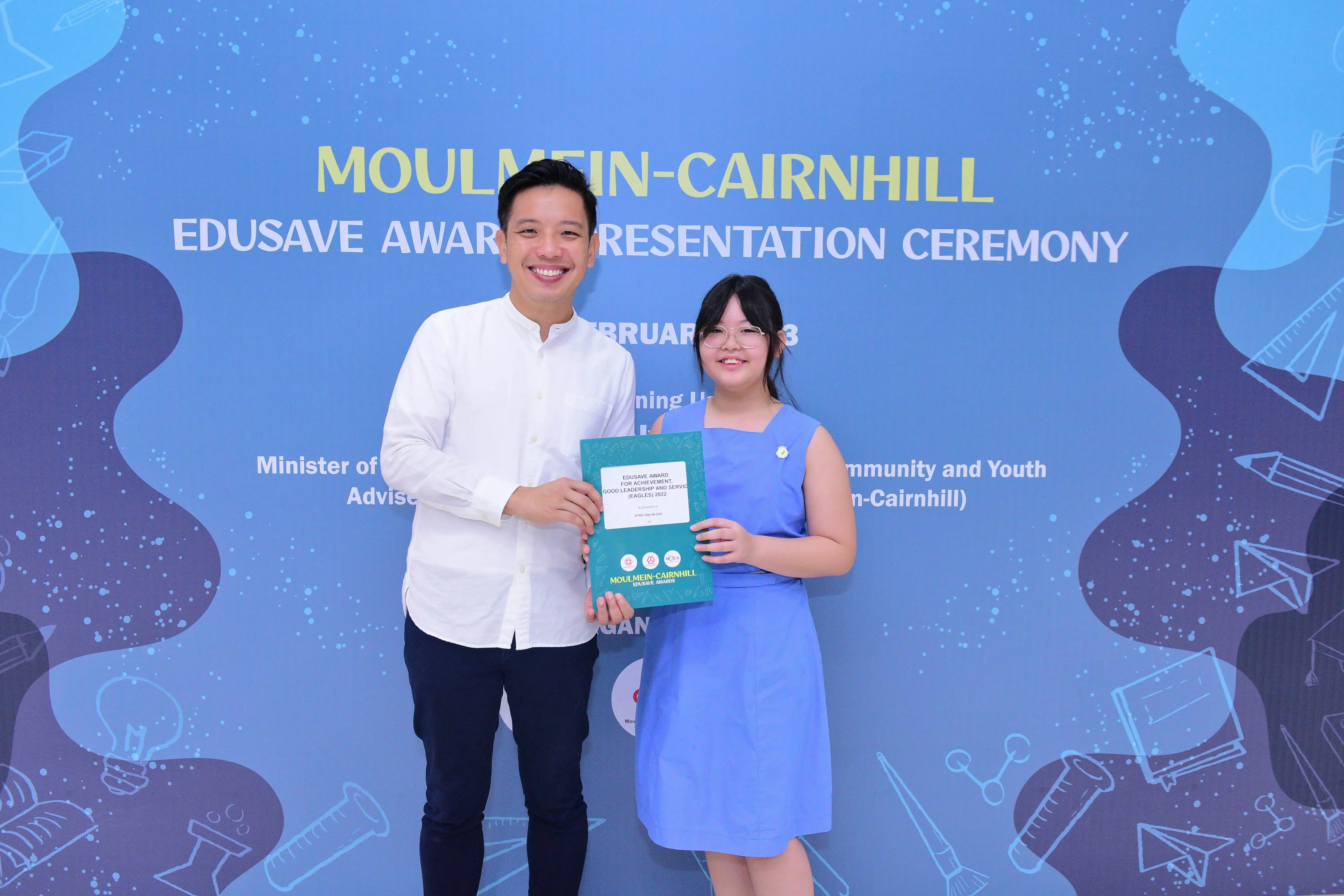 Two people smile holding a certificate at the Moulmein-Cairnhill Edusave Award Presentation Ceremony backdrop.