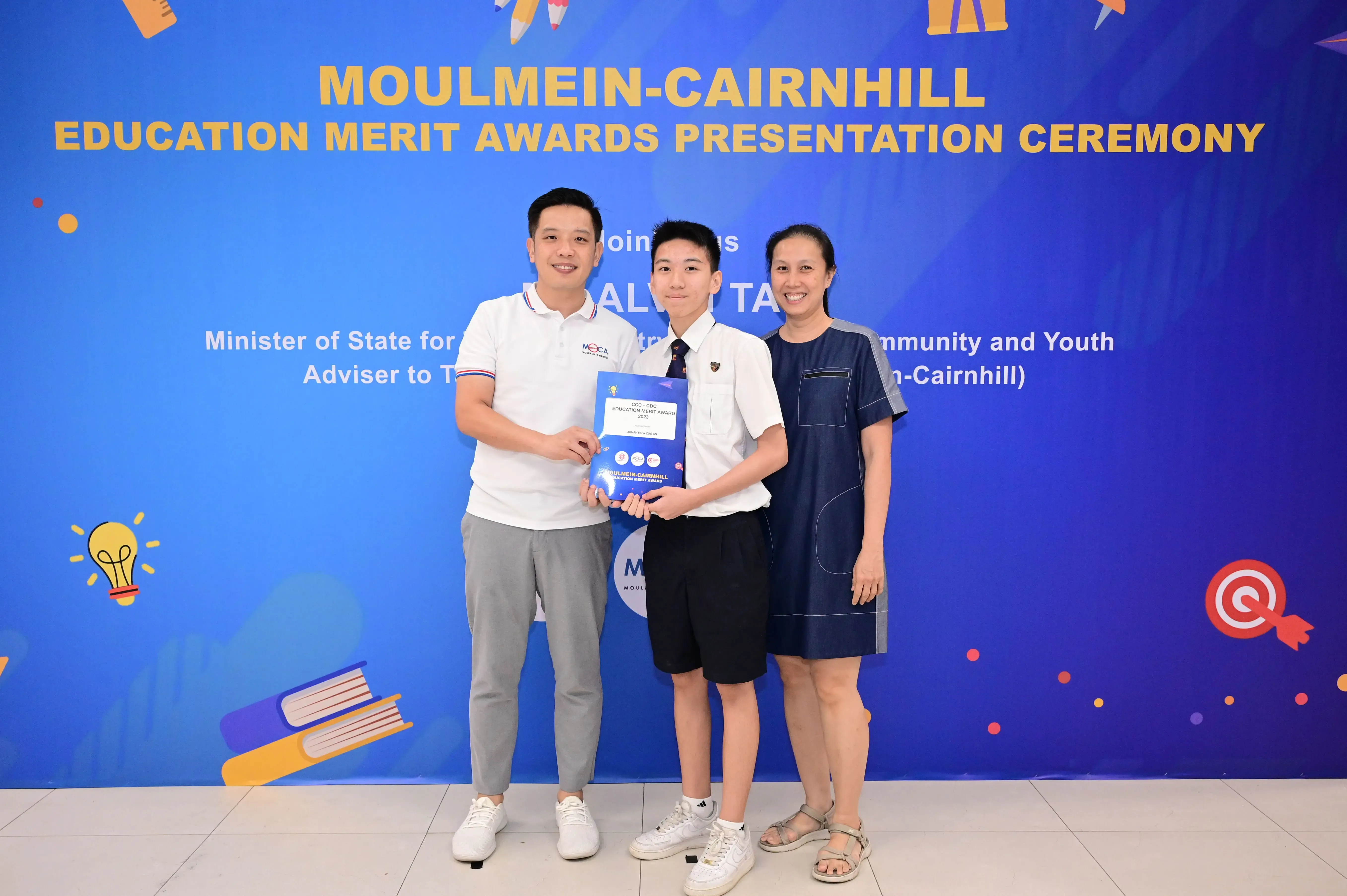 Three people pose holding an award booklet before a Moulmein-Cairnhill Education Merit Awards Presentation Ceremony banner.