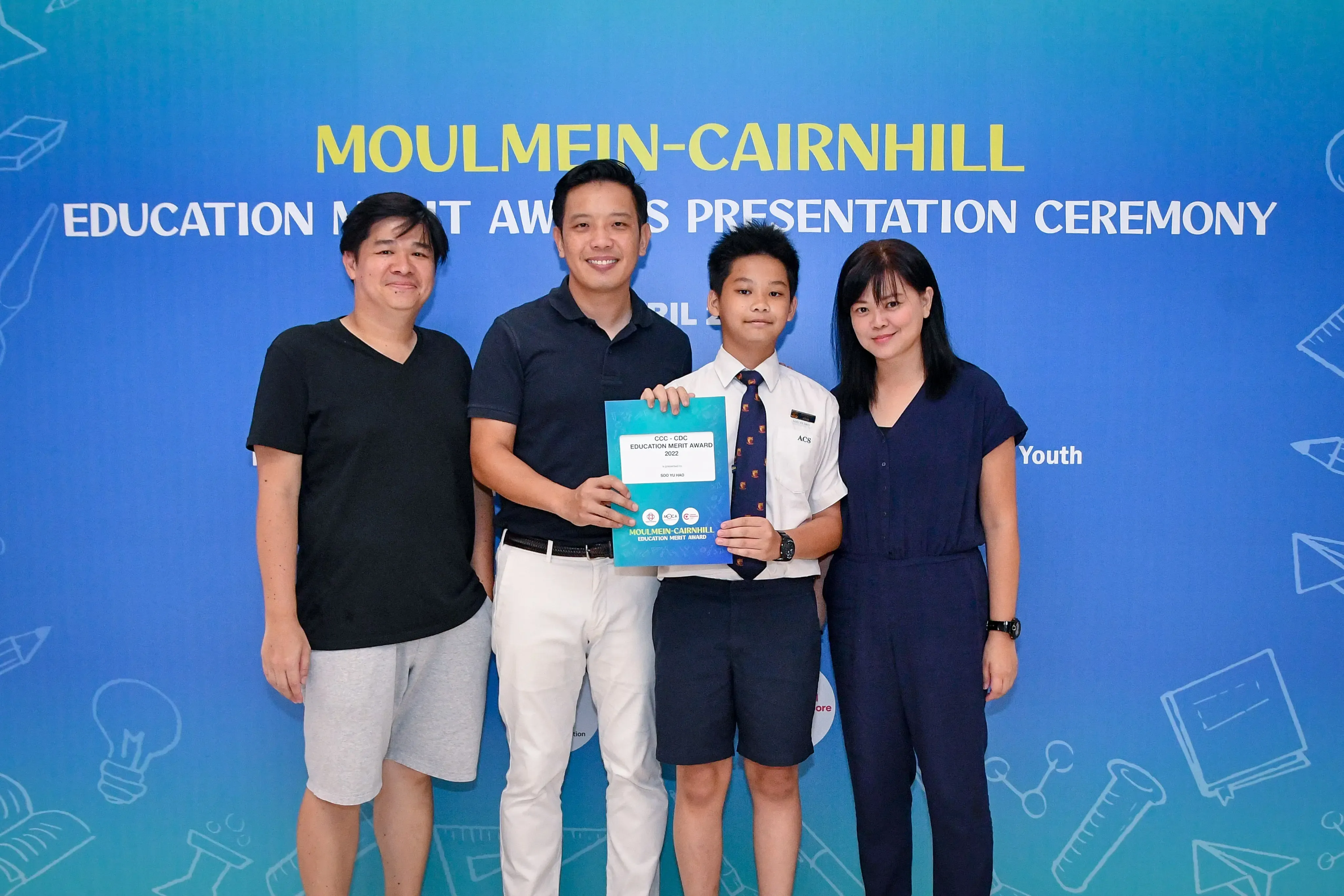 Four people pose at the Moulmein-Cairnhill Education Merit Awards Presentation Ceremony, with a boy holding a certificate.