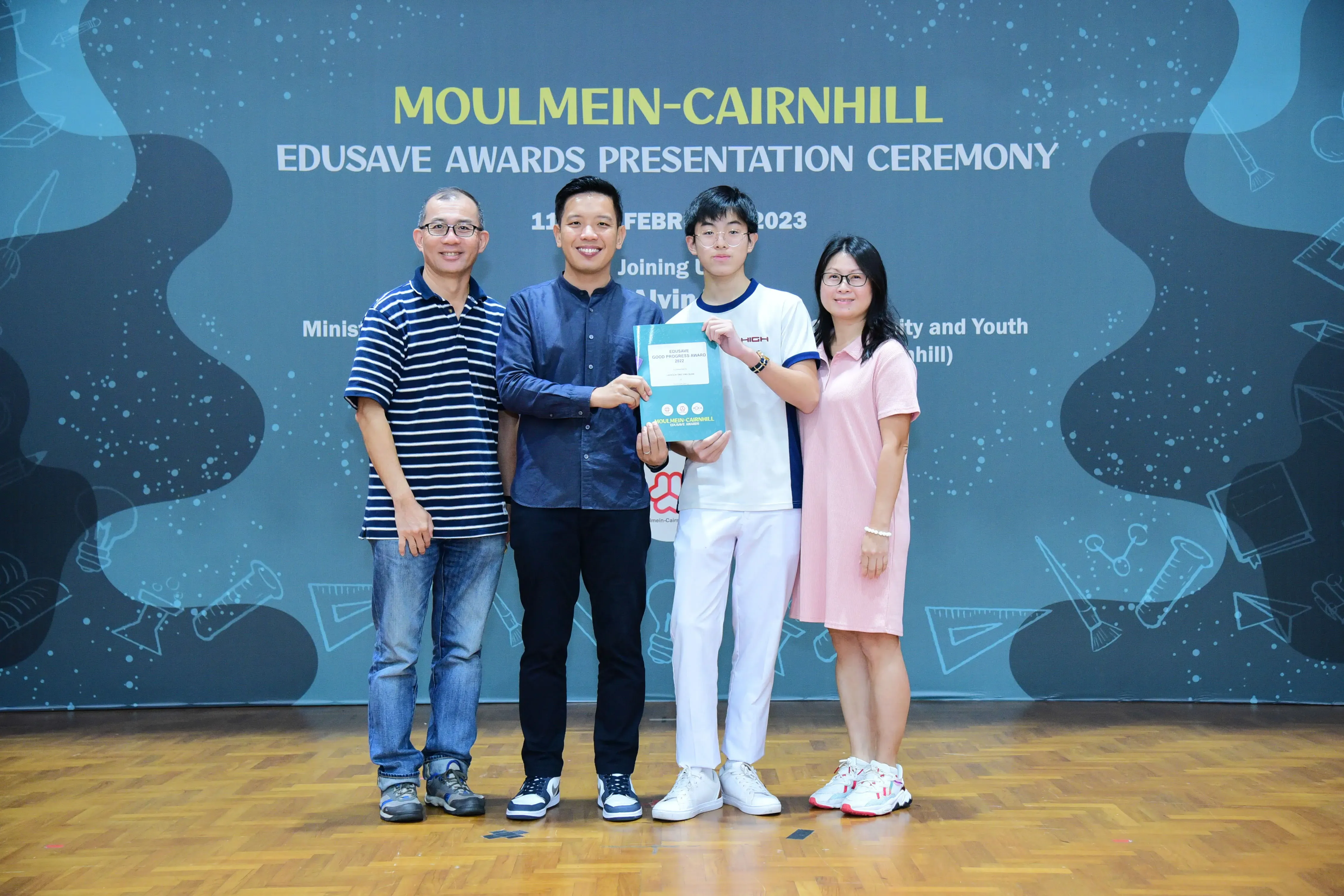 Four people pose on a stage at the Moulmein-Cairnhill Edusave Awards Presentation Ceremony, holding a certificate.