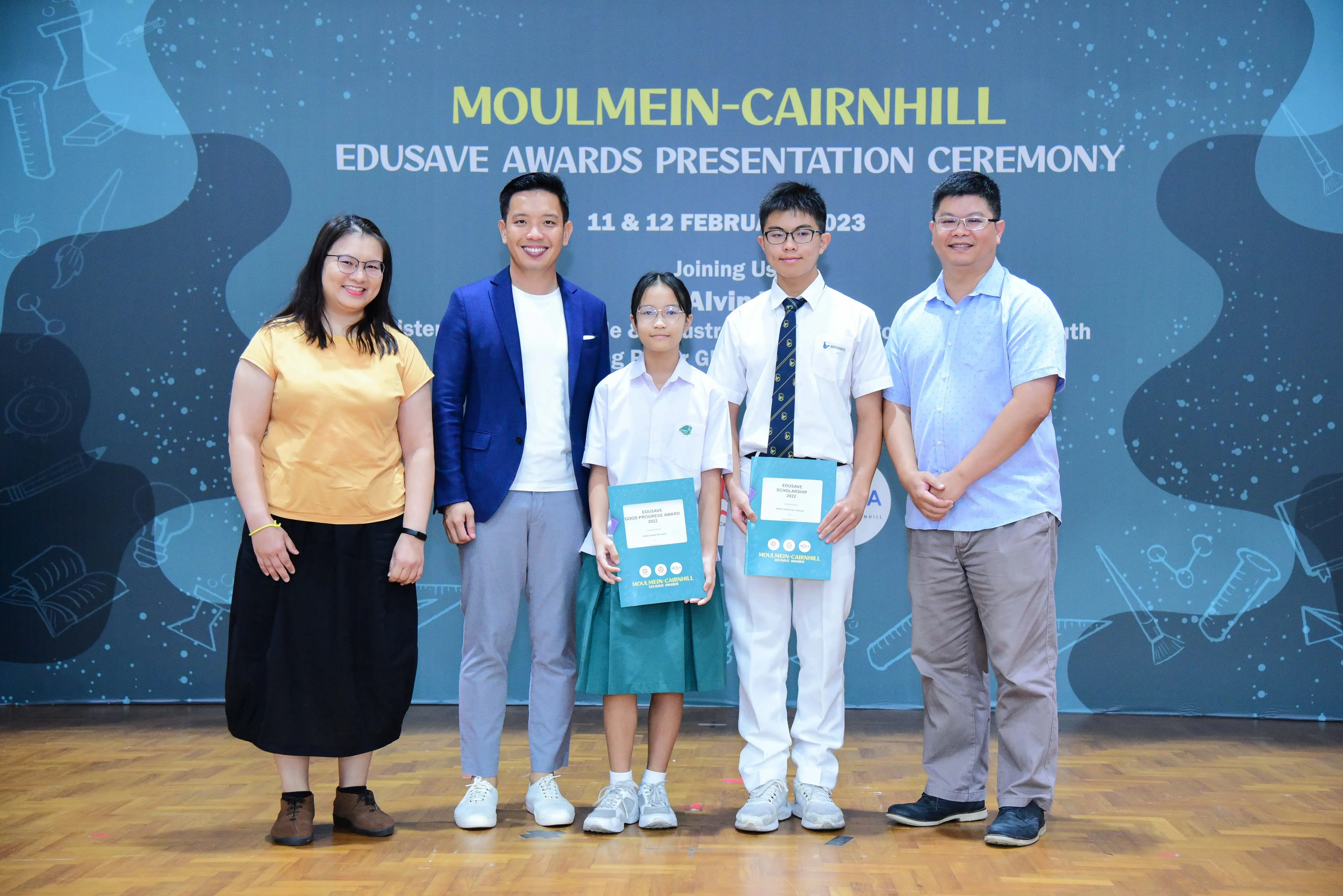 Five people pose at the Moulmein-Cairnhill Edusave Awards Presentation Ceremony, two students holding teal award folders.