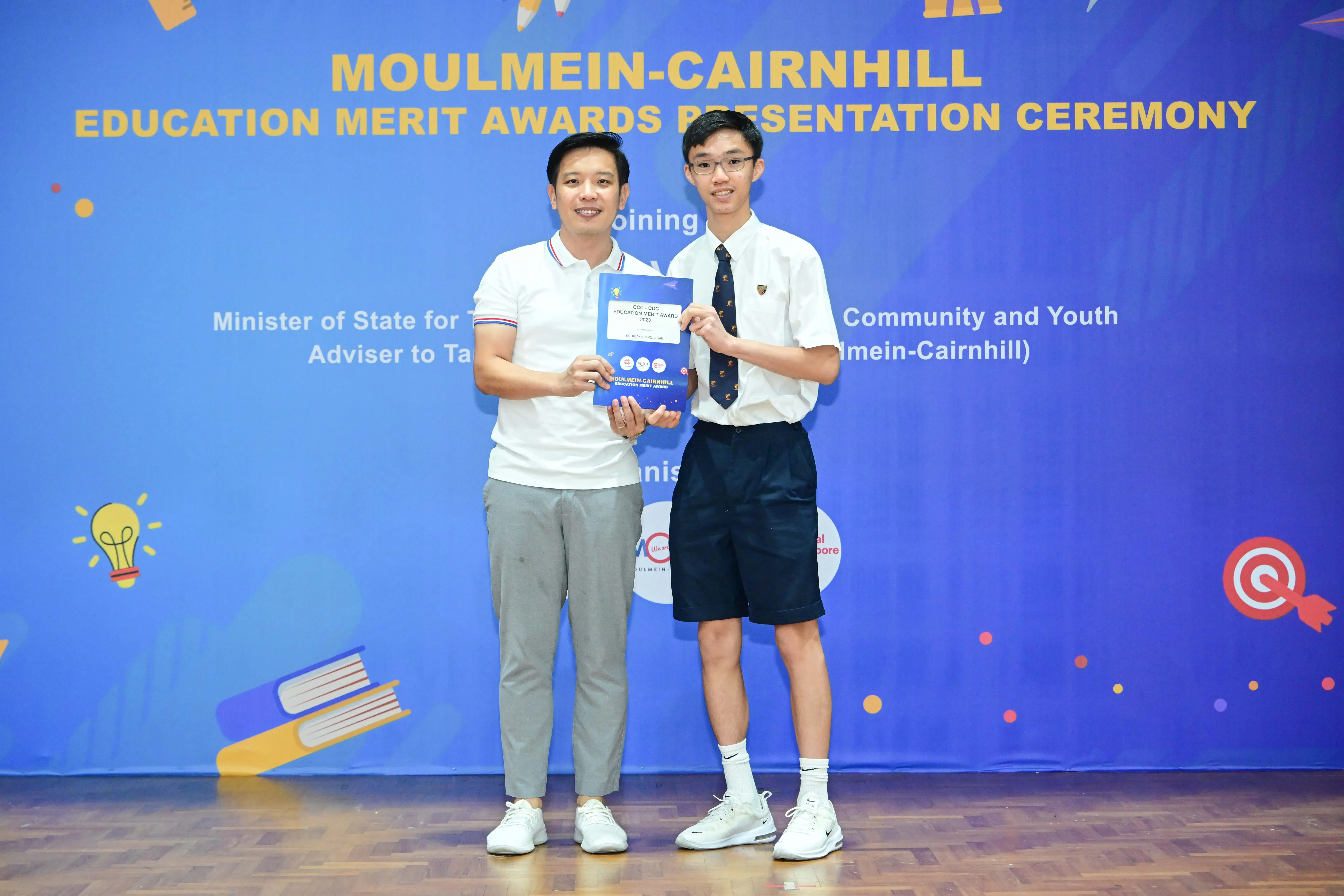 Two men hold a blue booklet at the Moulmein-Cairnhill Education Merit Awards Presentation Ceremony backdrop.