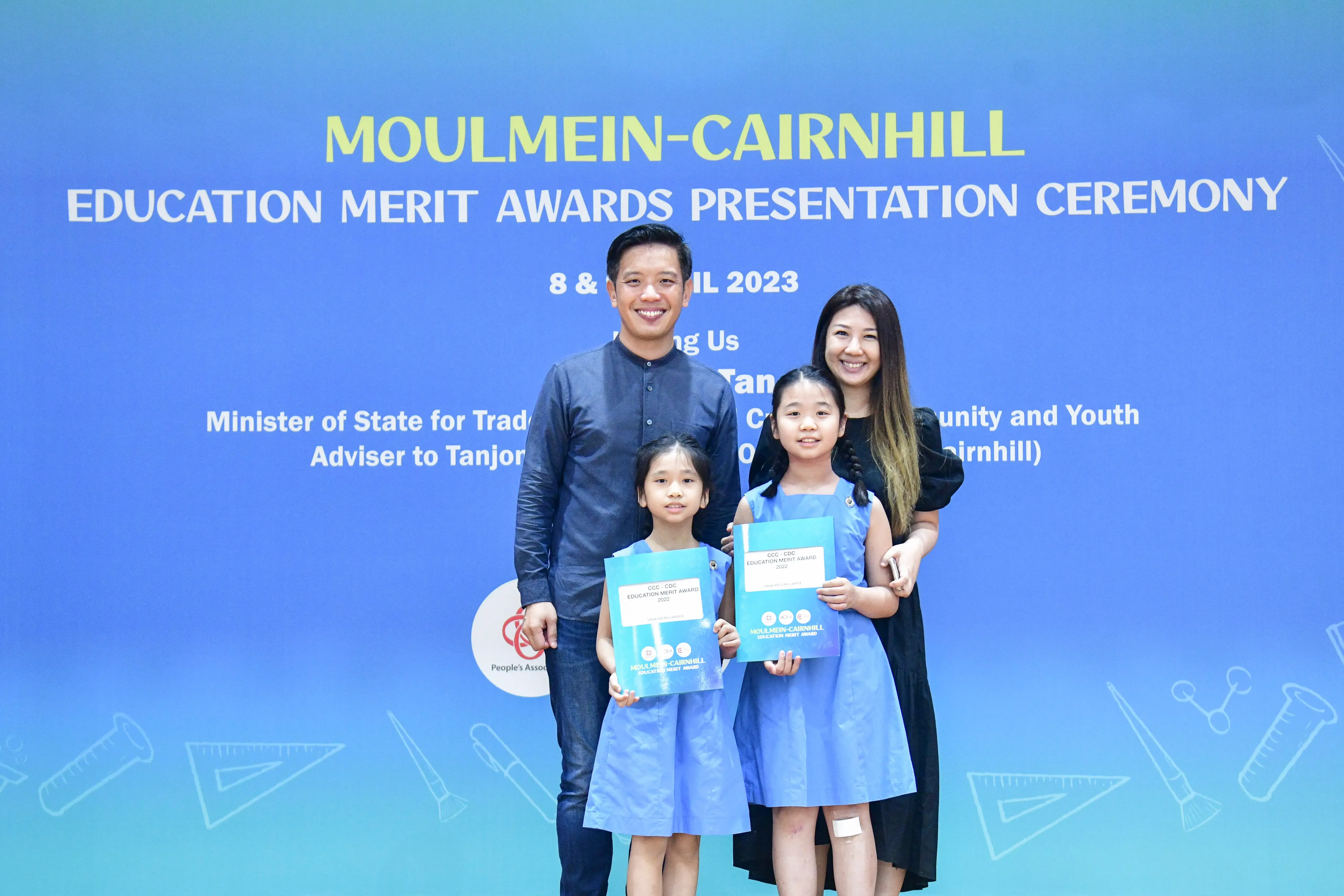 Two adults and two girls holding award folders pose before a Moulmein-Cairnhill Education Merit Awards 2023 banner.