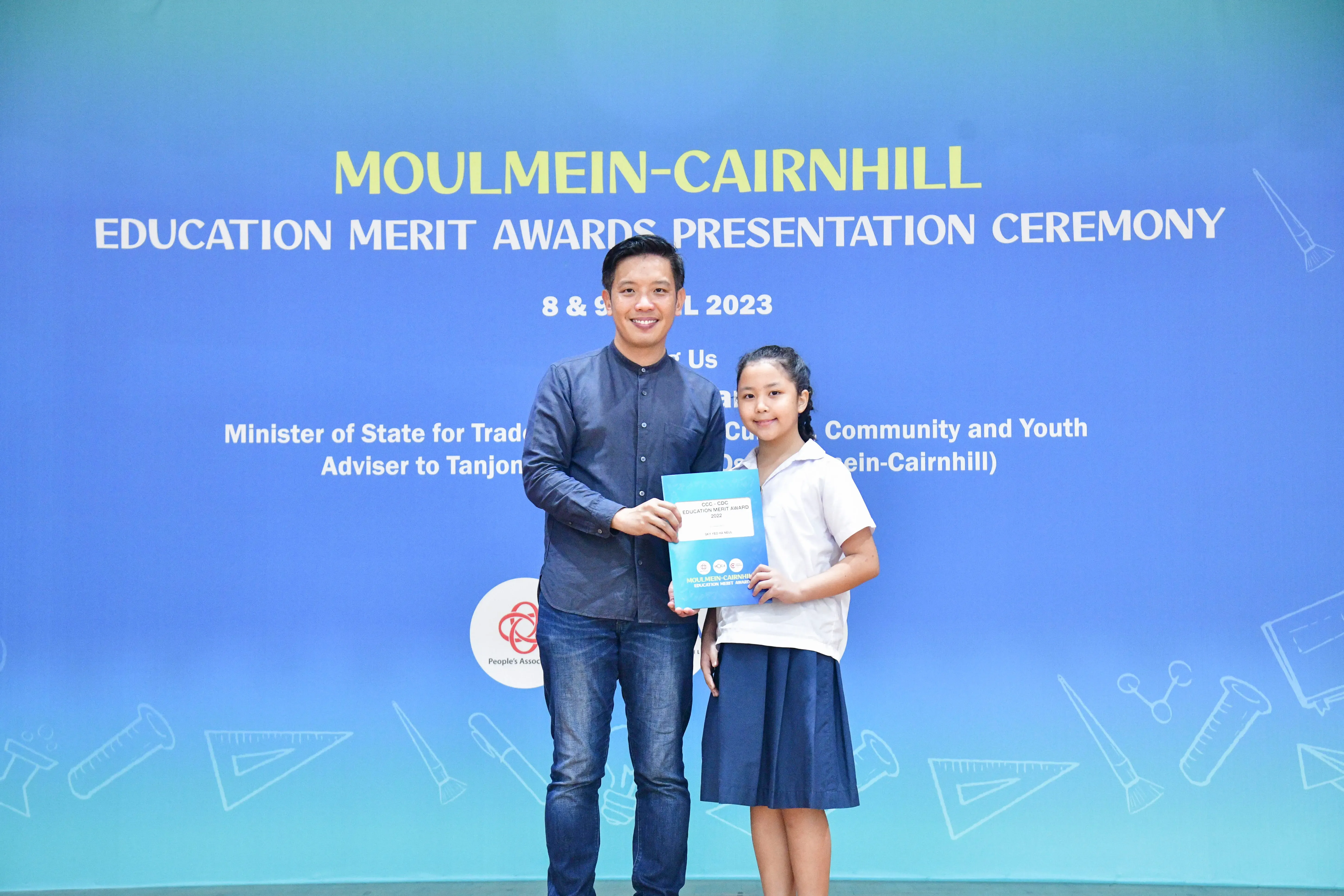 A man and a young girl hold a certificate together in front of a Moulmein-Cairnhill Education Merit Awards 2023 banner.