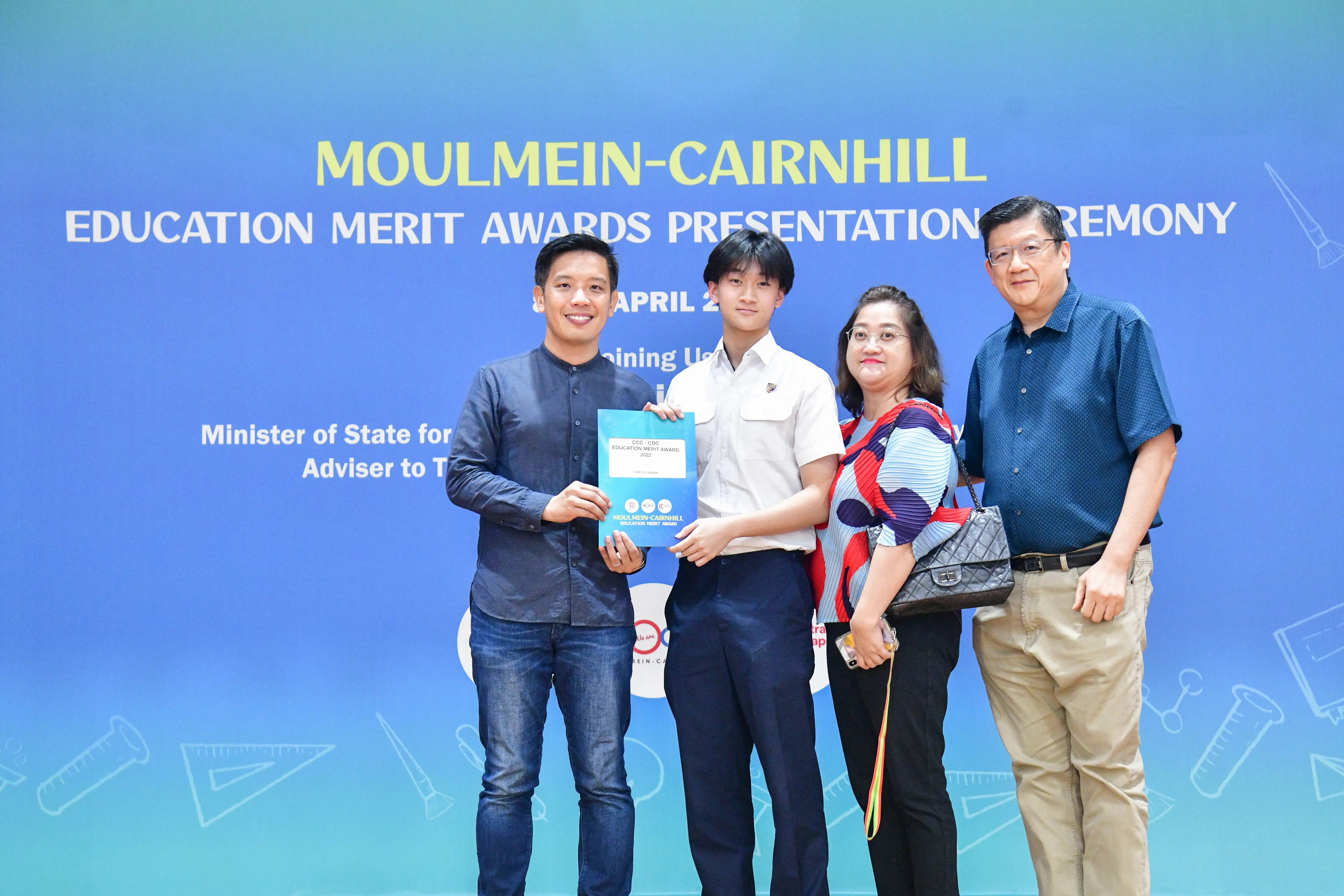 Four people pose at the Moulmein-Cairnhill Education Merit Awards ceremony, with a young student holding a certificate.