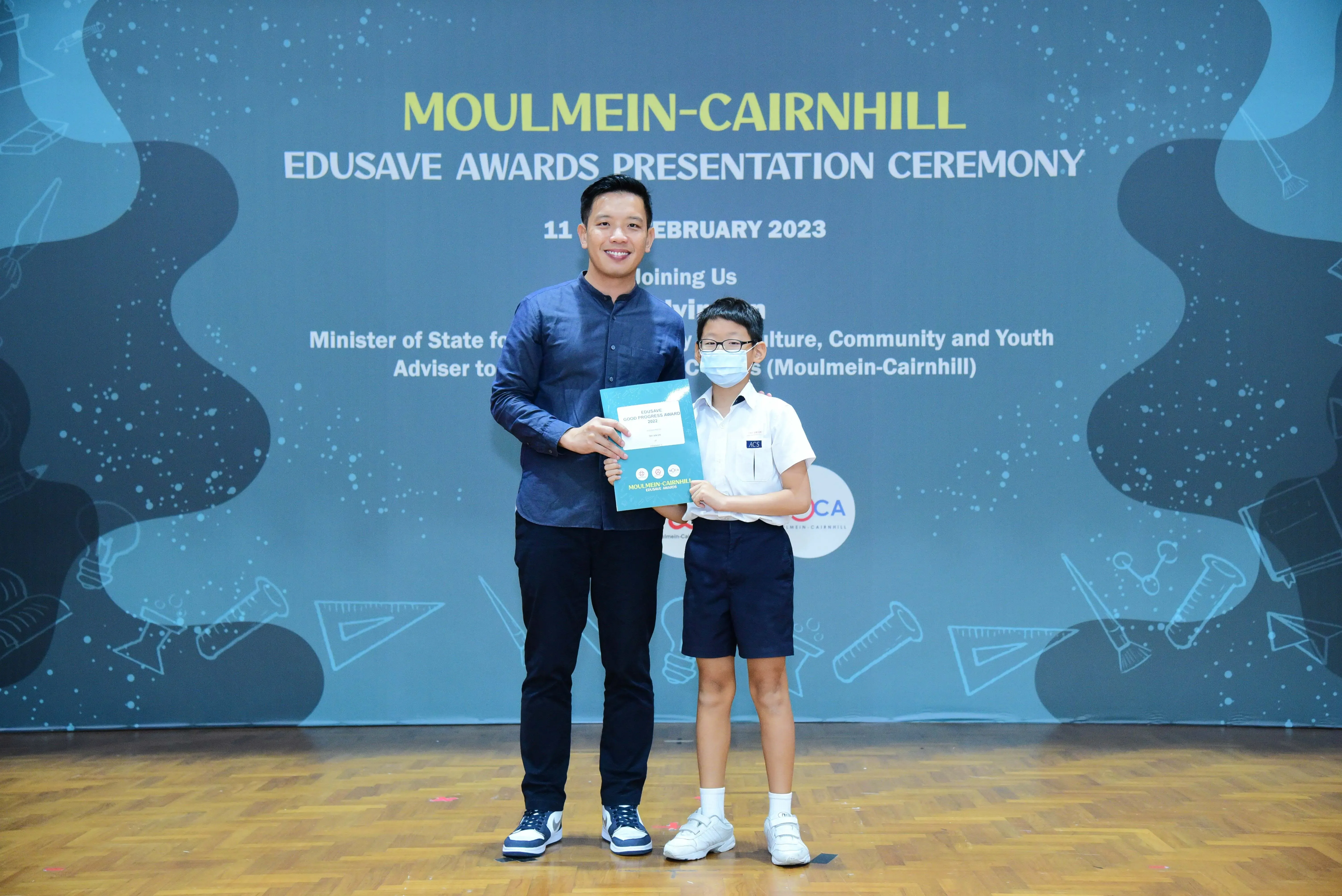 An adult and a young student pose with a certificate at the Moulmein-Cairnhill Edusave Awards Ceremony, February 2023.