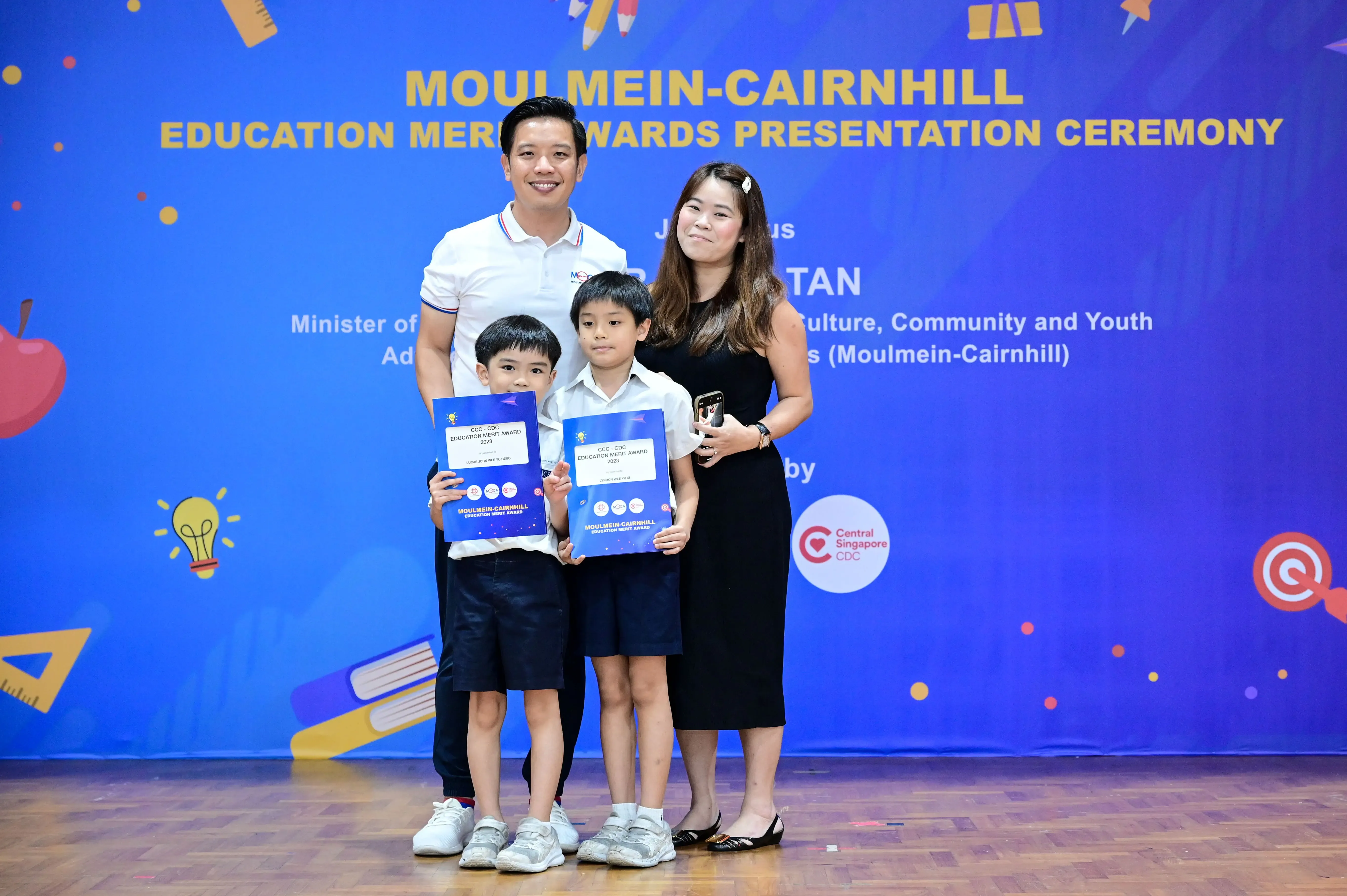 Two adults and two boys holding blue award certificates pose at the Moulmein-Cairnhill Education Merit Awards ceremony.
