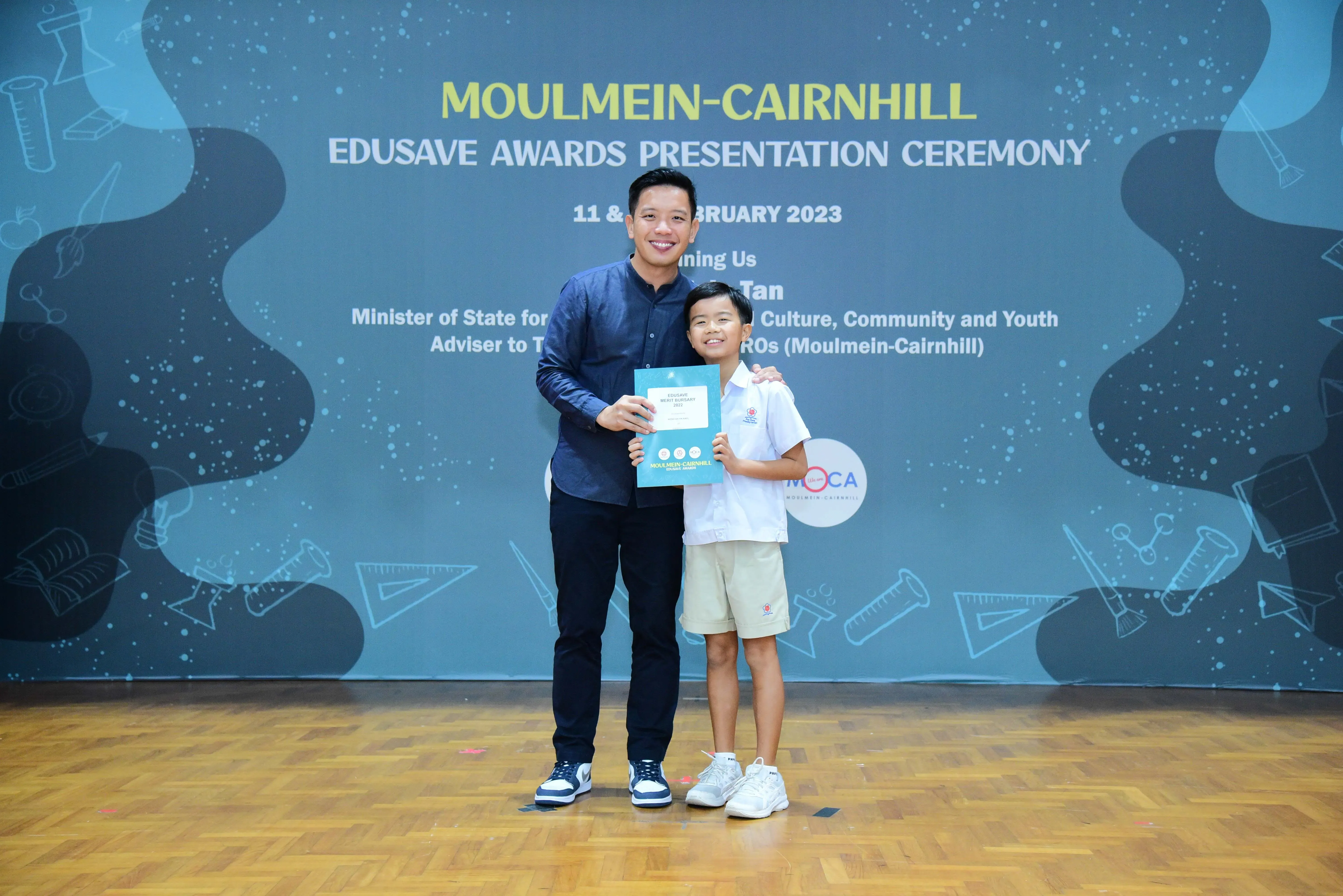 An adult and a young student hold a certificate at the Moulmein-Cairnhill Edusave Awards Ceremony 2023.