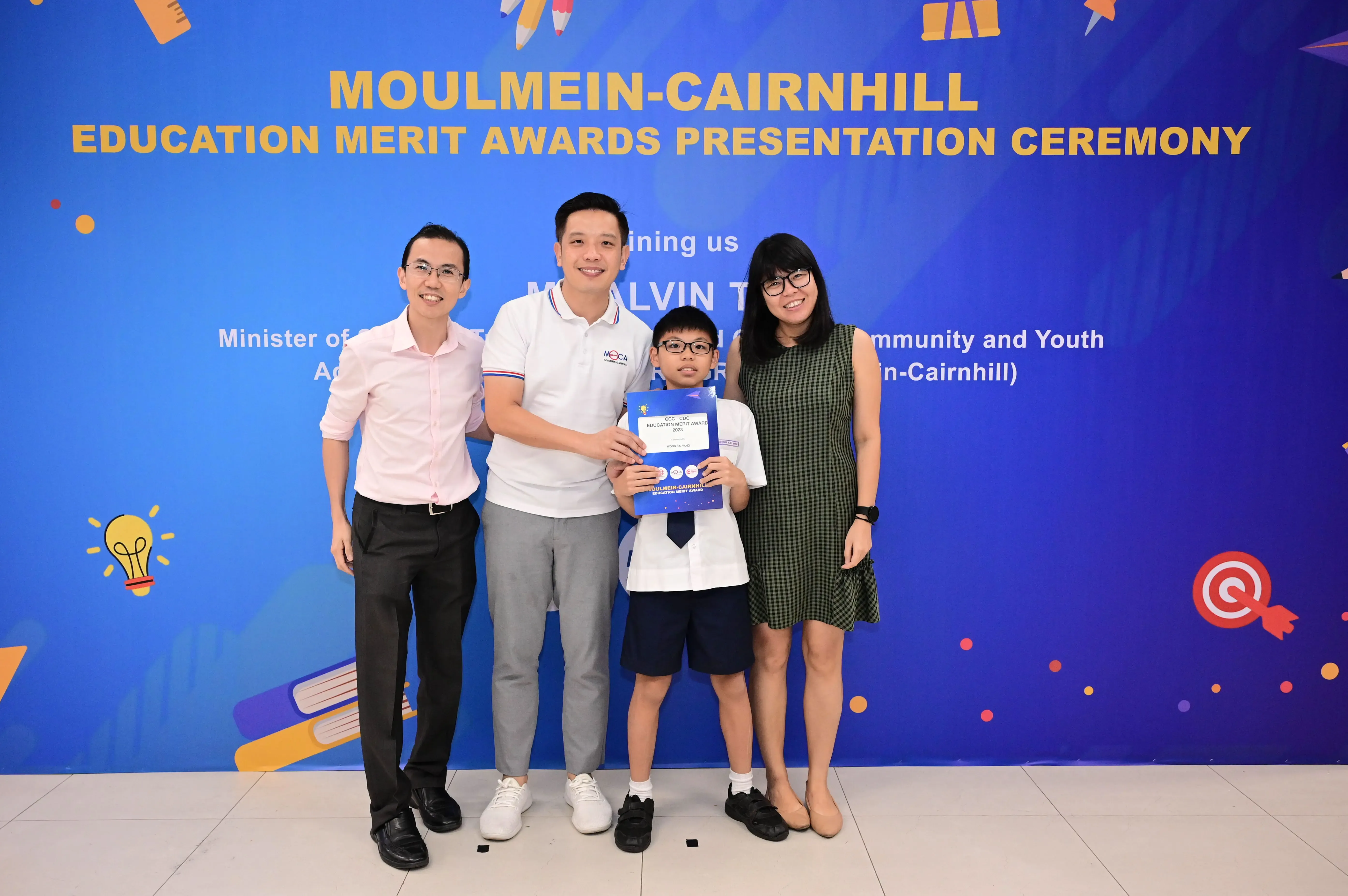 Four people pose before a Moulmein-Cairnhill Education Merit Awards banner, with a boy holding a blue certificate.