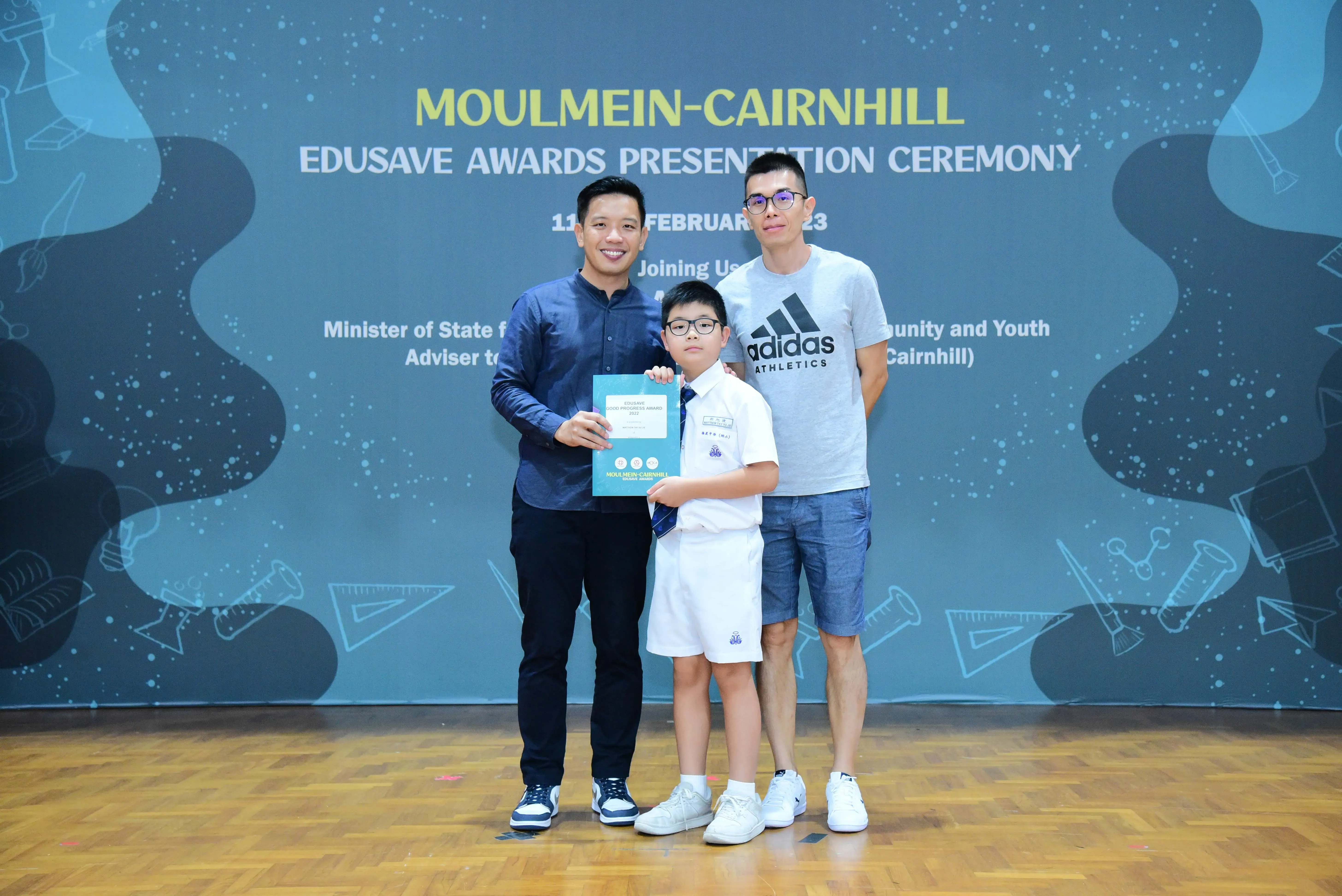 Two men and a young boy in school uniform hold a certificate at the Moulmein-Cairnhill Edusave Awards Ceremony.