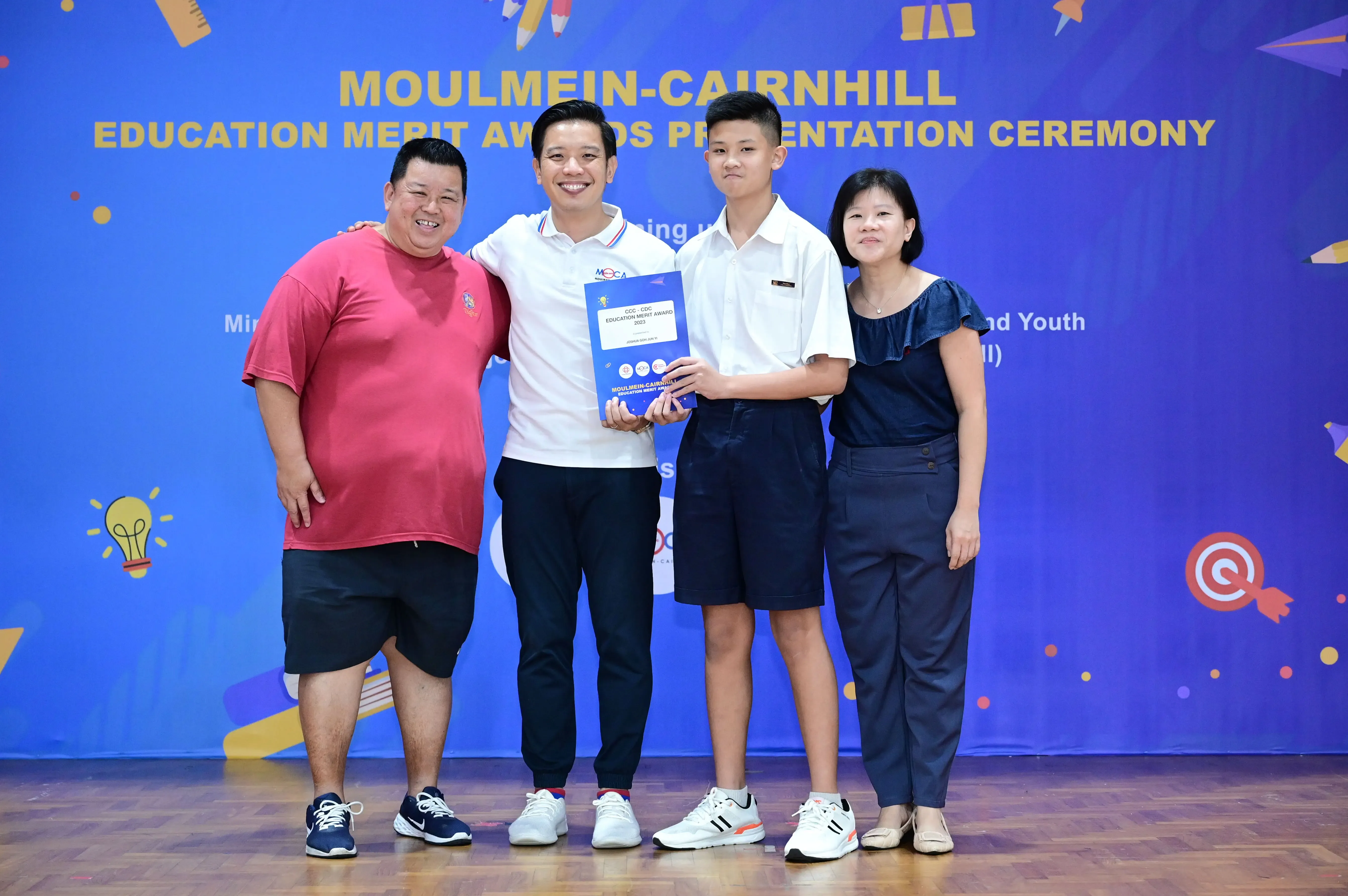 Four people pose on stage at the Moulmein-Cairnhill Education Merit Awards Presentation Ceremony, holding an award.
