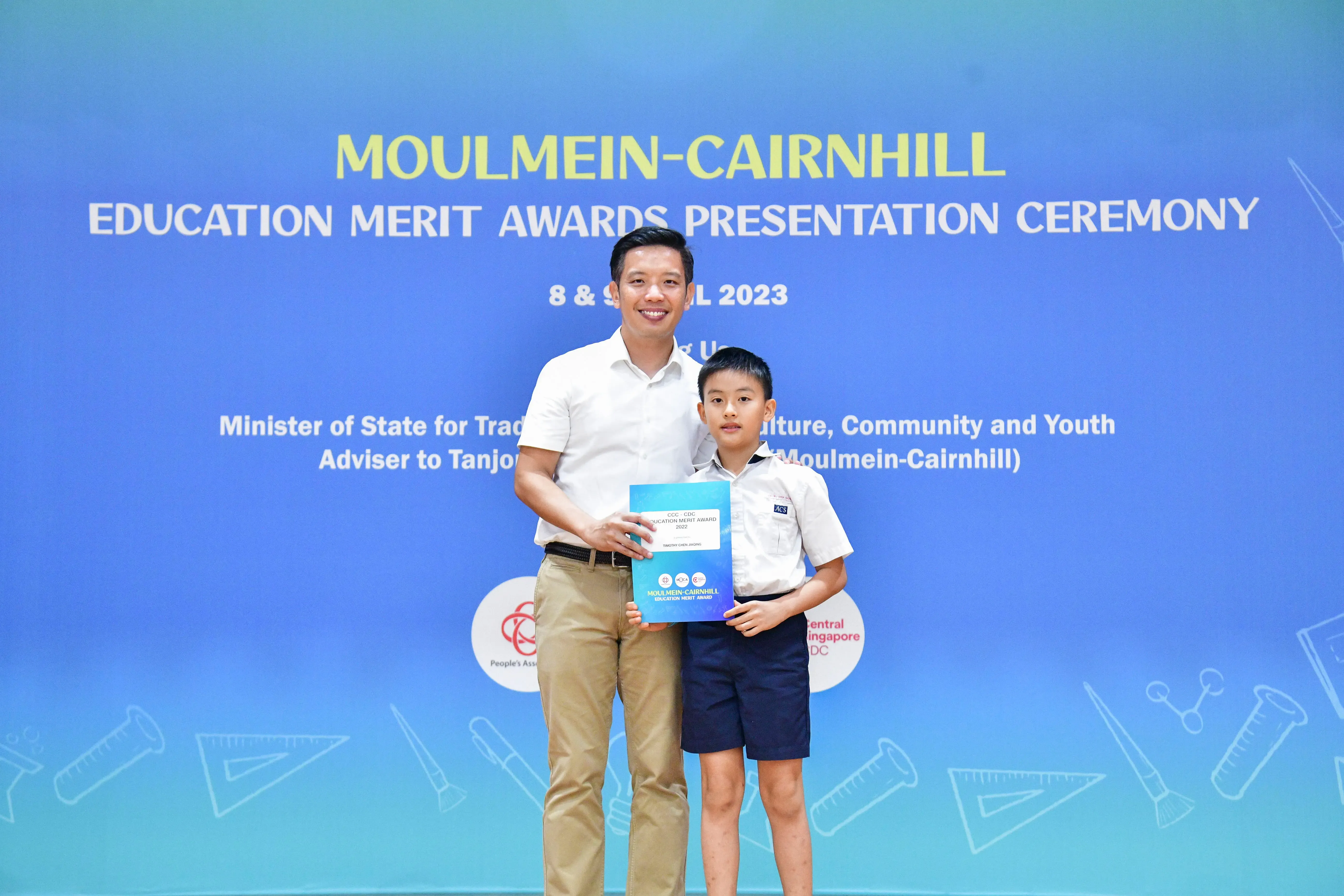 An adult man and a young boy hold an award certificate at the Moulmein-Cairnhill Education Merit Awards Ceremony 2023.