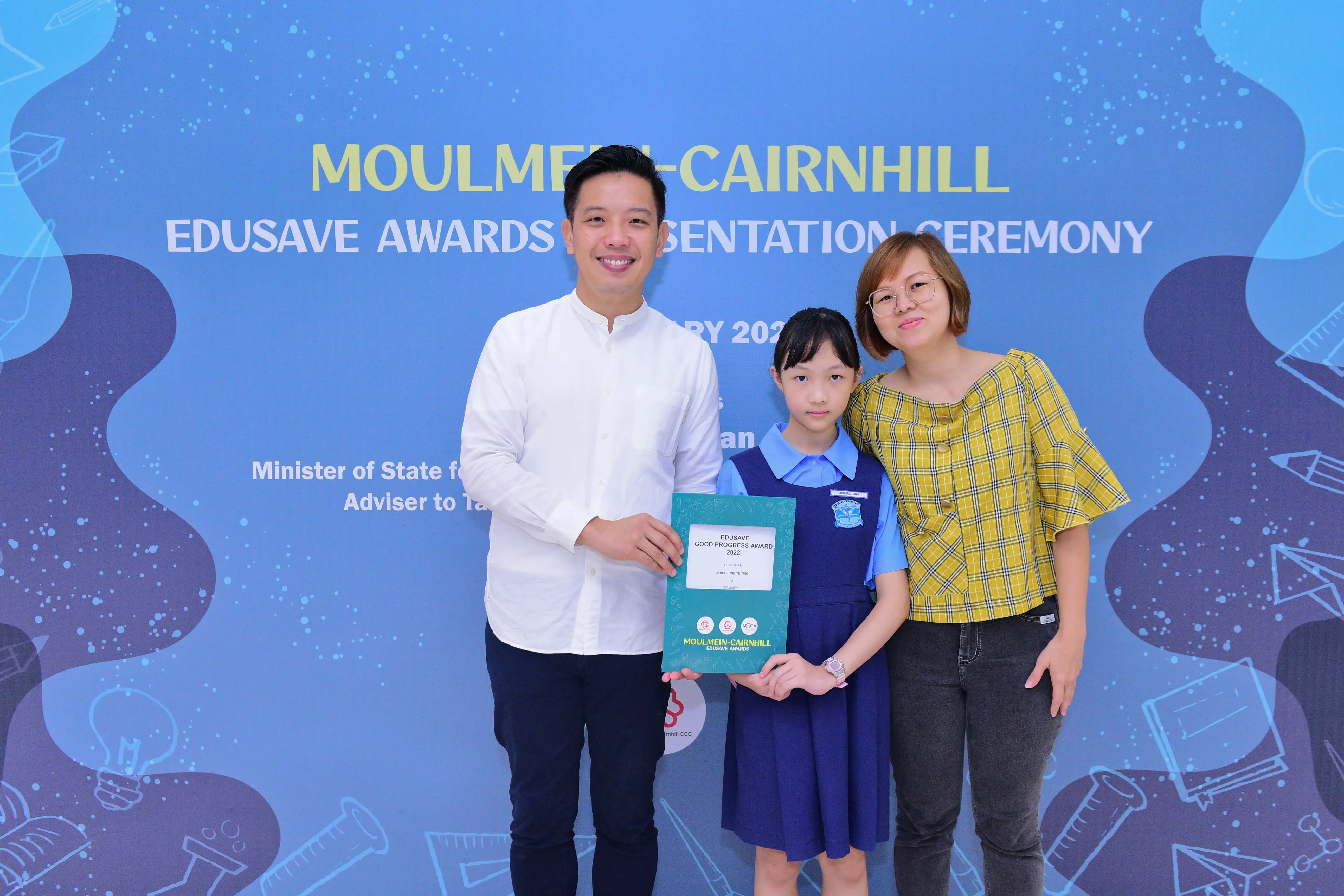 A girl in school uniform holds an Edusave award certificate flanked by a man and woman at an awards ceremony.