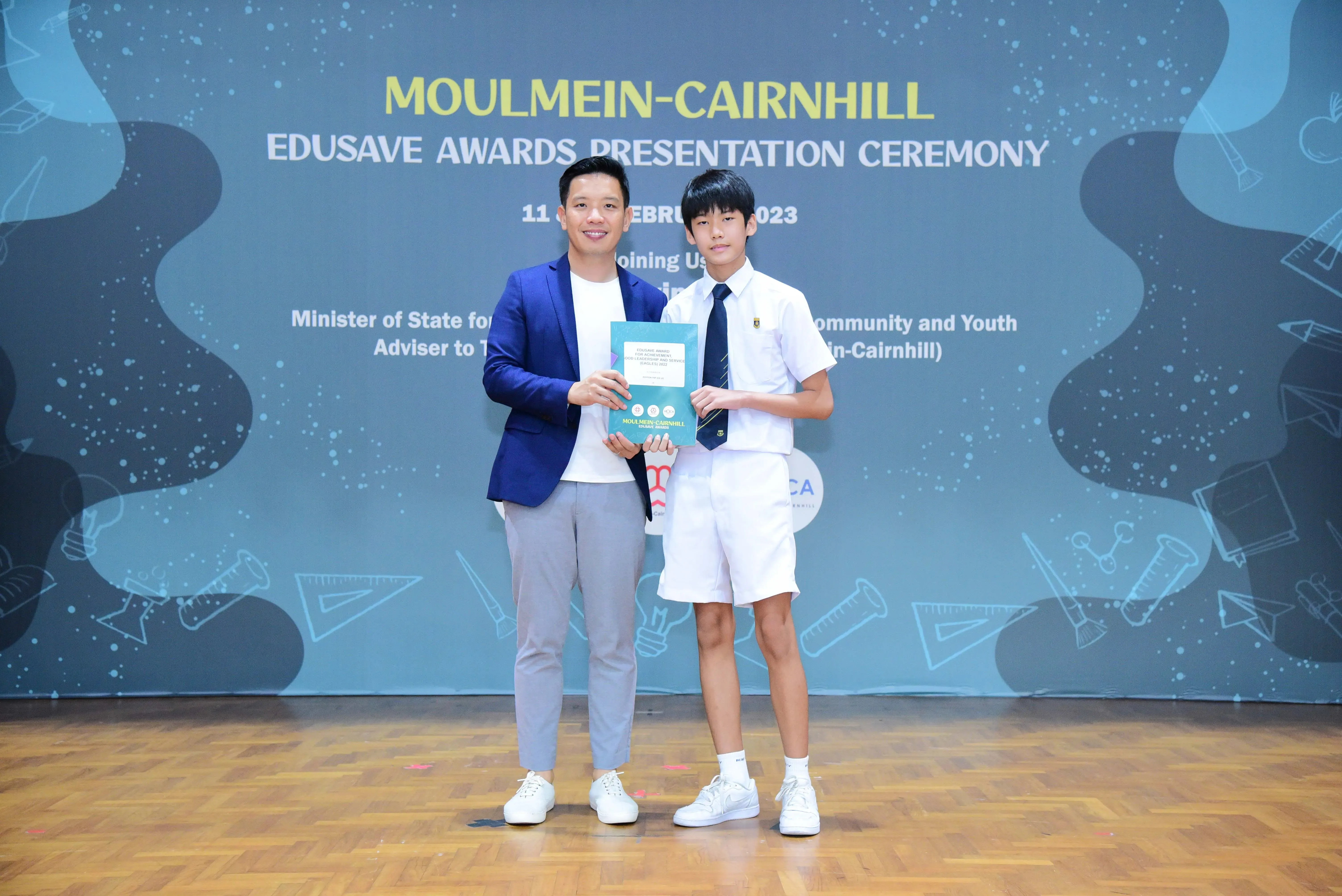 An adult in a blue blazer and a student in school uniform hold an award at the Moulmein-Cairnhill Edusave Awards Ceremony 2023.
