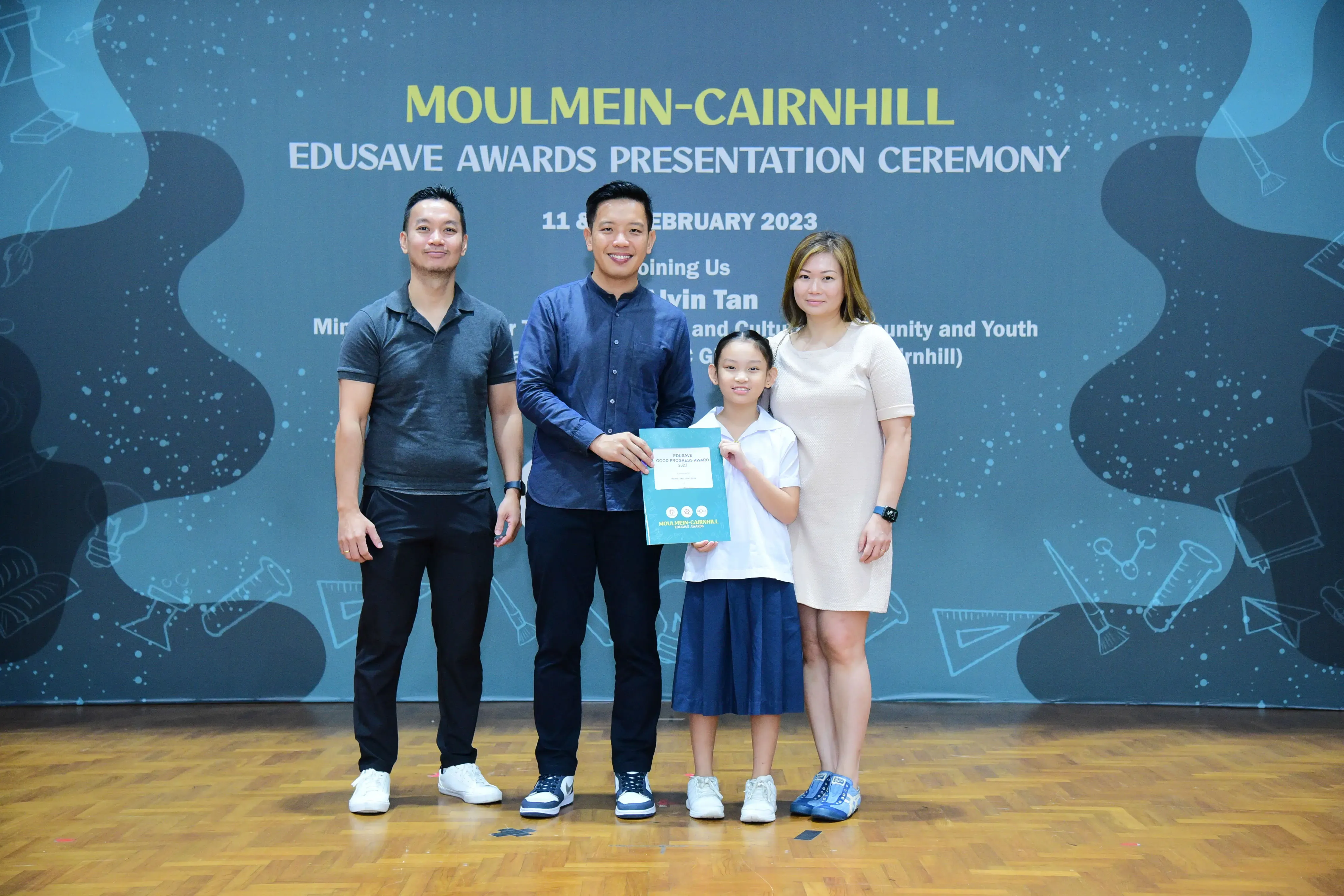 Four people pose at the Moulmein-Cairnhill Edusave Awards Presentation Ceremony, with a young girl holding a certificate.