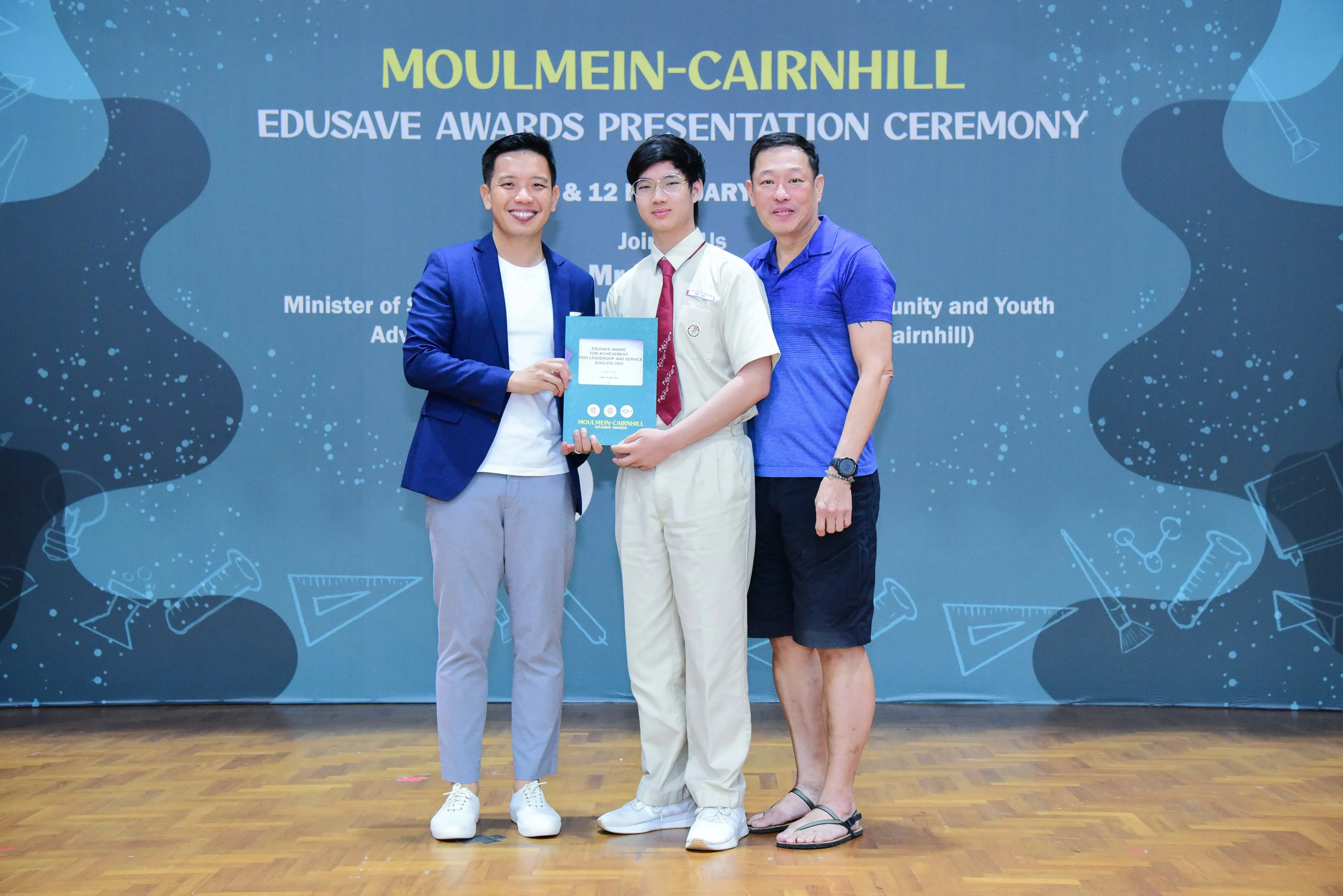Three people pose at the Moulmein-Cairnhill Edusave Awards Presentation Ceremony, with a student holding an award booklet.