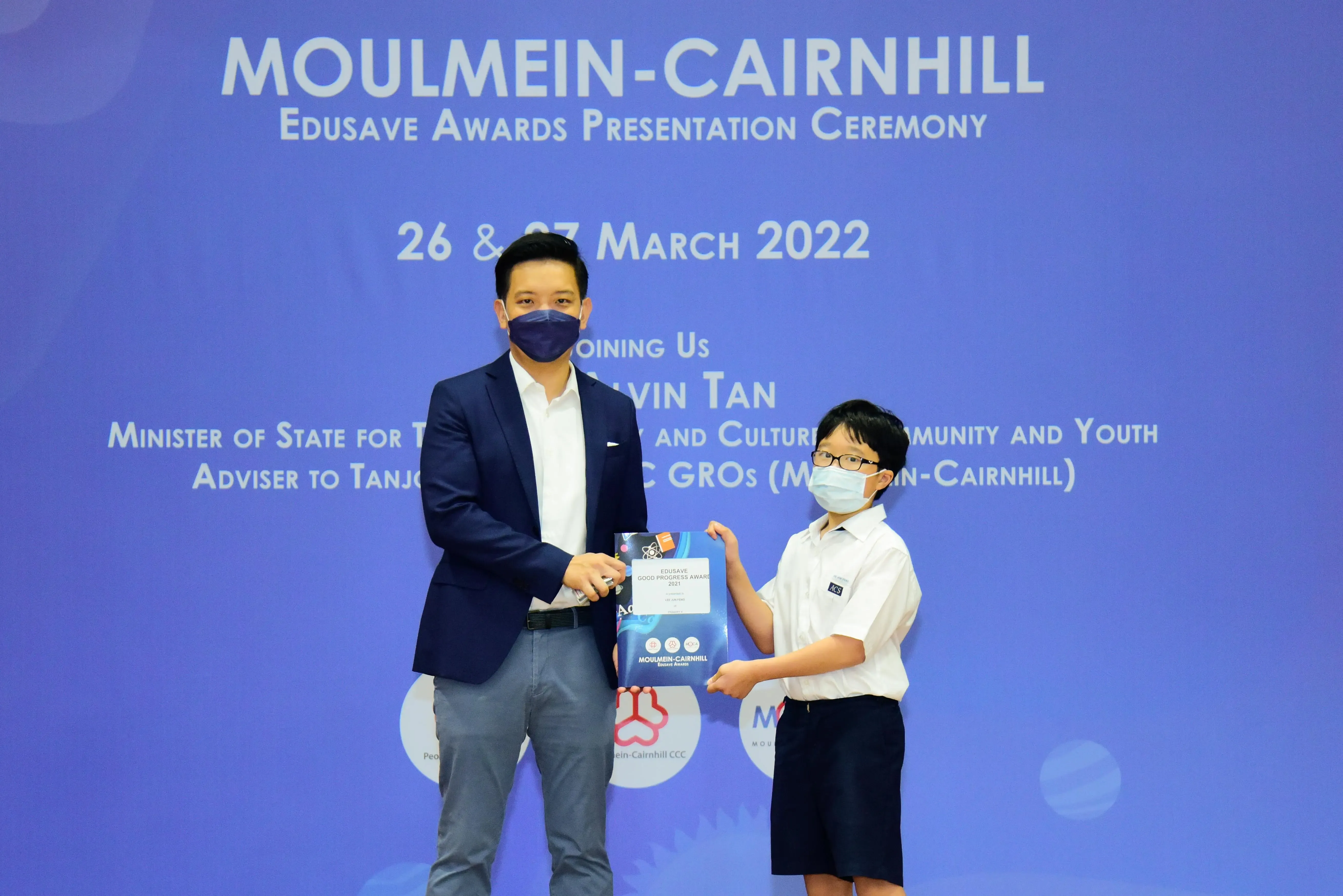 Adult in a suit and a student in uniform exchange an award package at the Moulmein-Cairnhill Edusave ceremony, March 2022.