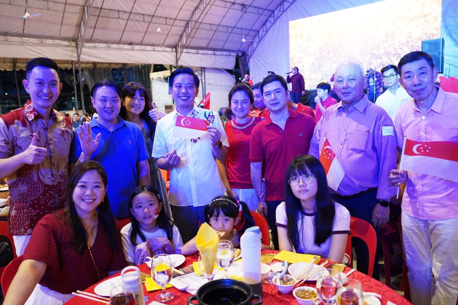 Group of people smiling at a festive event, holding small Singapore flags, seated and standing around a dining table.