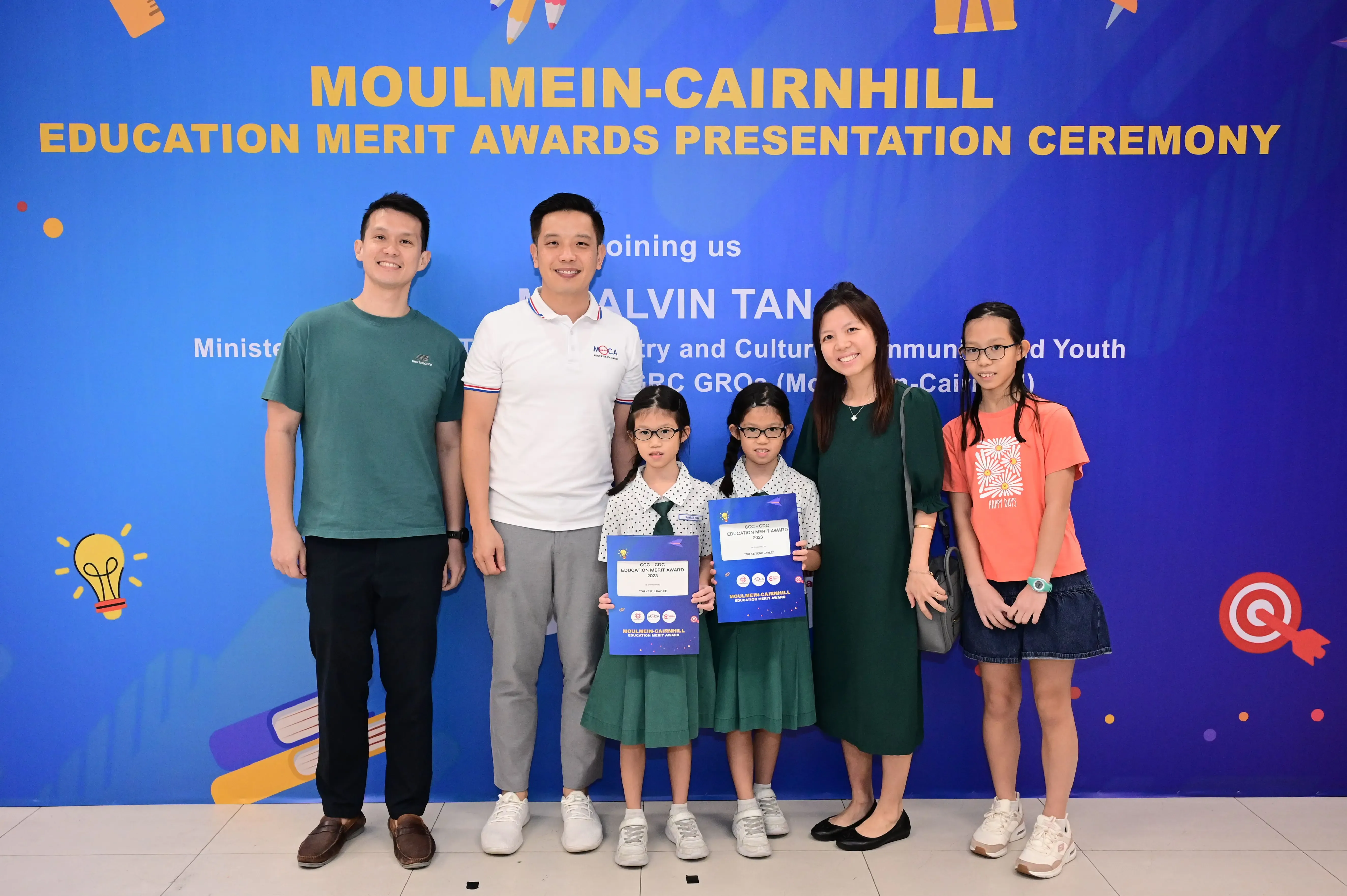 Six people pose before a blue Moulmein-Cairnhill Education Merit Awards banner, two young girls holding award certificates.