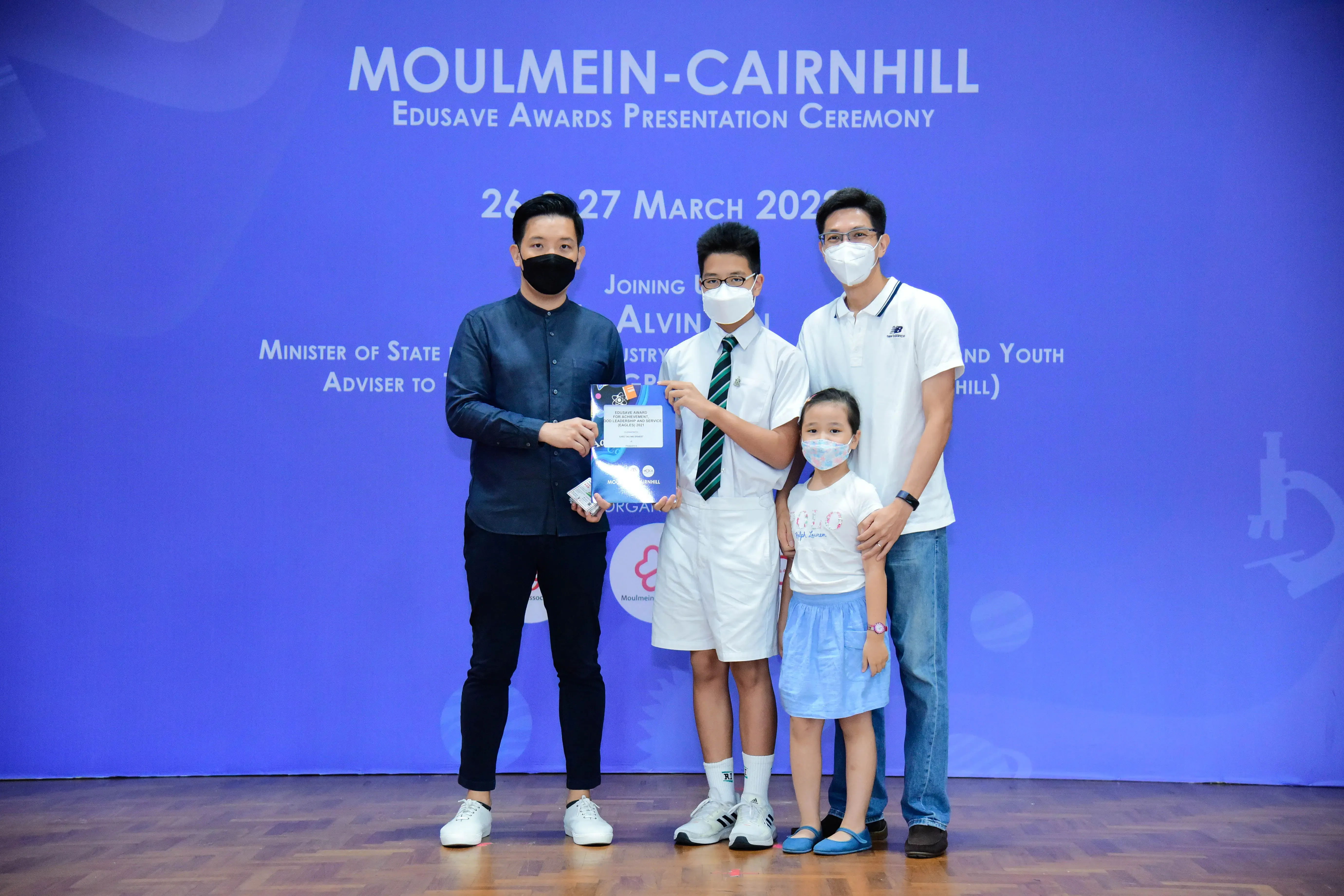 Four masked people pose at the Moulmein-Cairnhill Edusave Awards Presentation Ceremony, with a student holding a certificate.