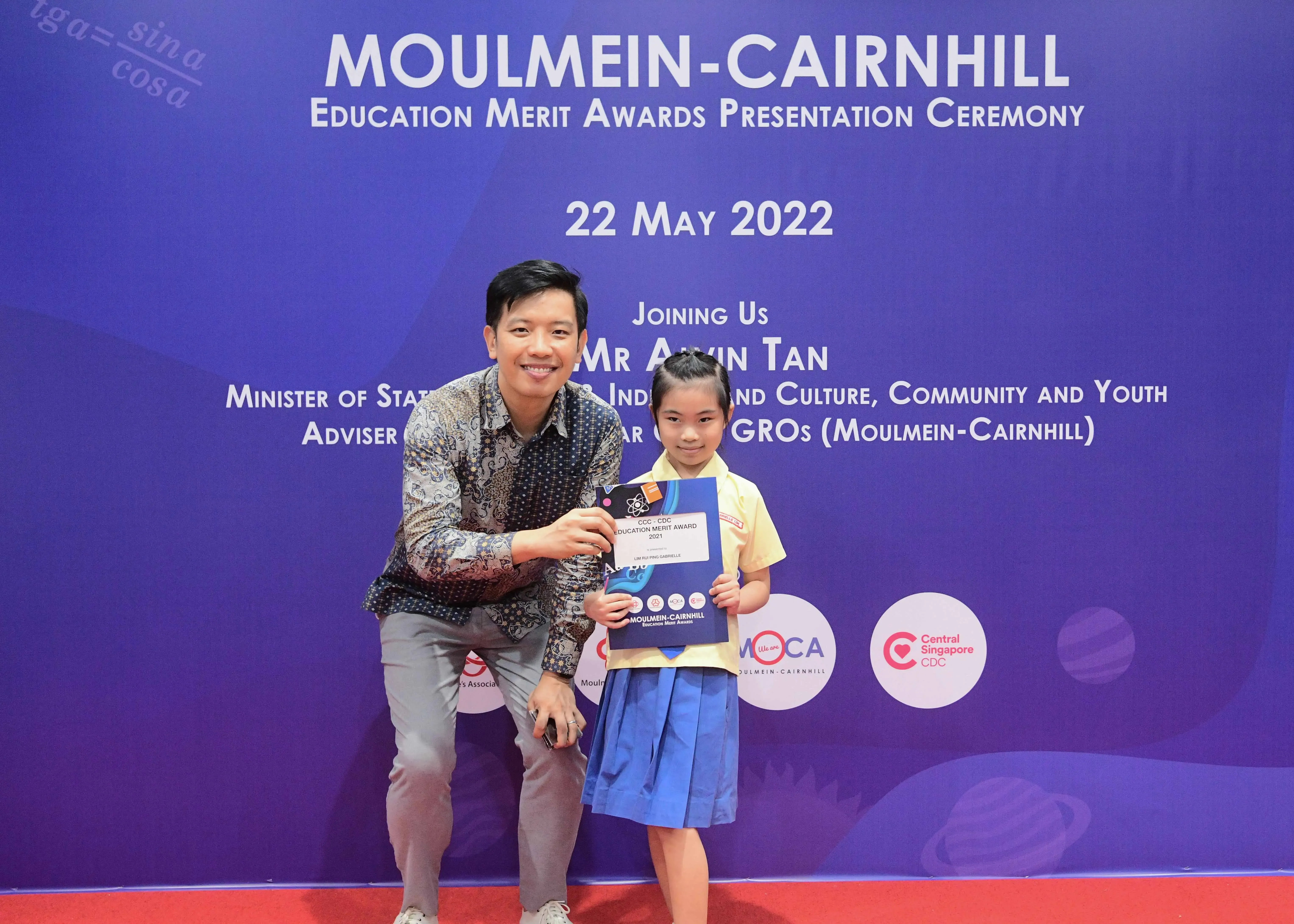 A man kneels beside a young girl holding a certificate at the Moulmein-Cairnhill Education Merit Awards, 22 May 2022.