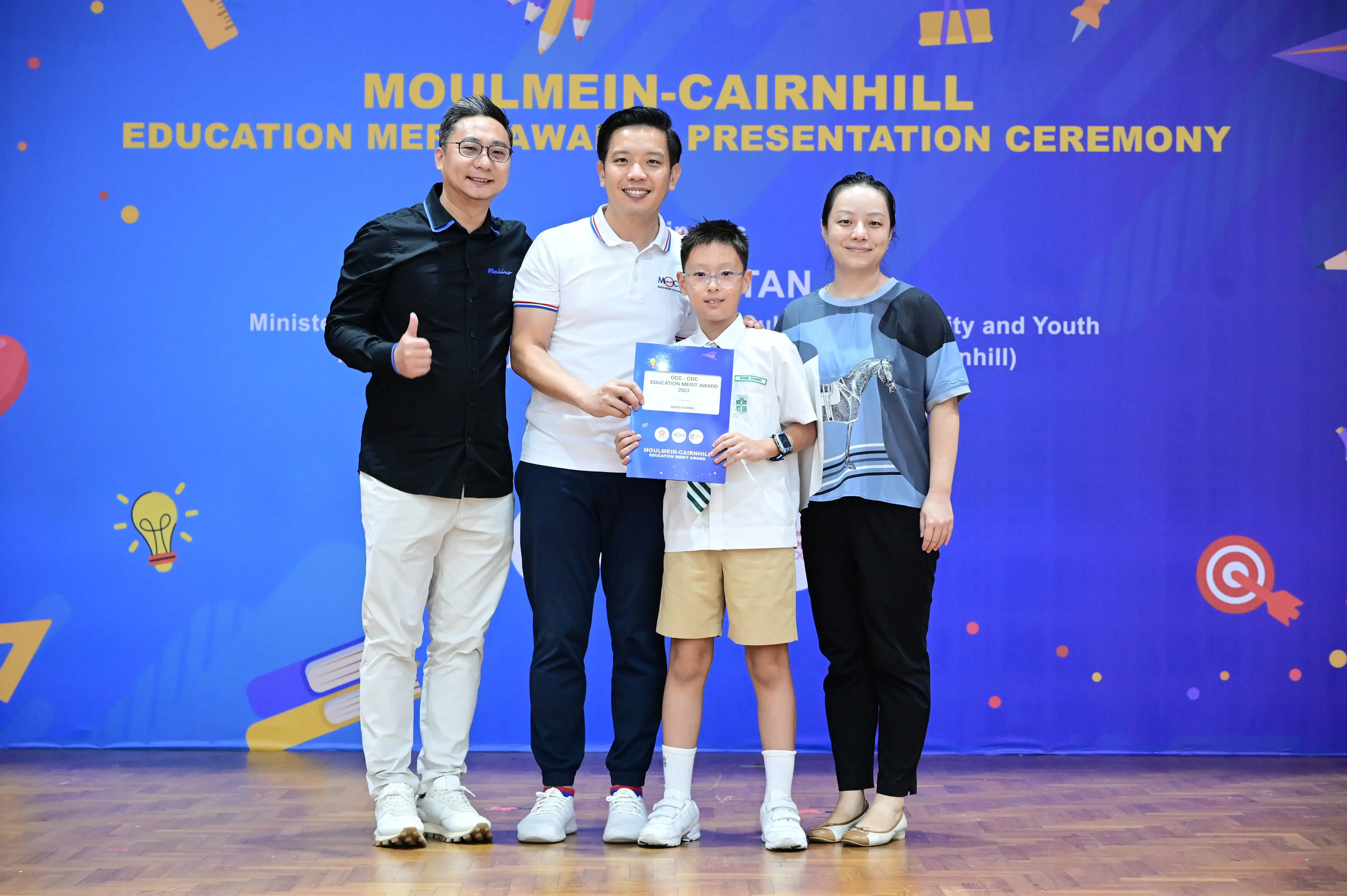 Three adults and a young boy pose holding an award certificate at the Moulmein-Cairnhill Education Merit Award ceremony.