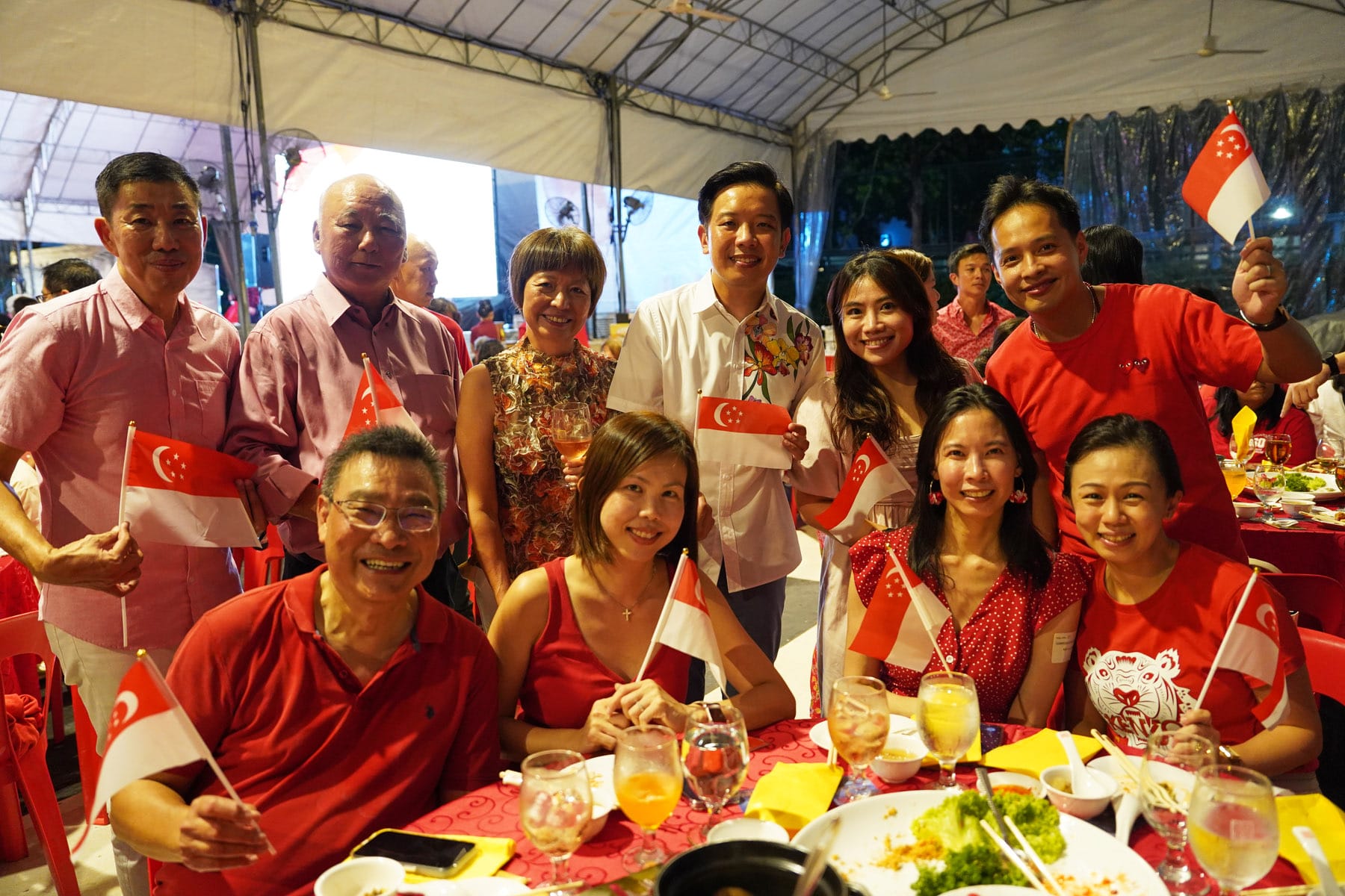 Group of people in red outfits smiling and holding Singapore flags at an outdoor dinner event under a tent.
