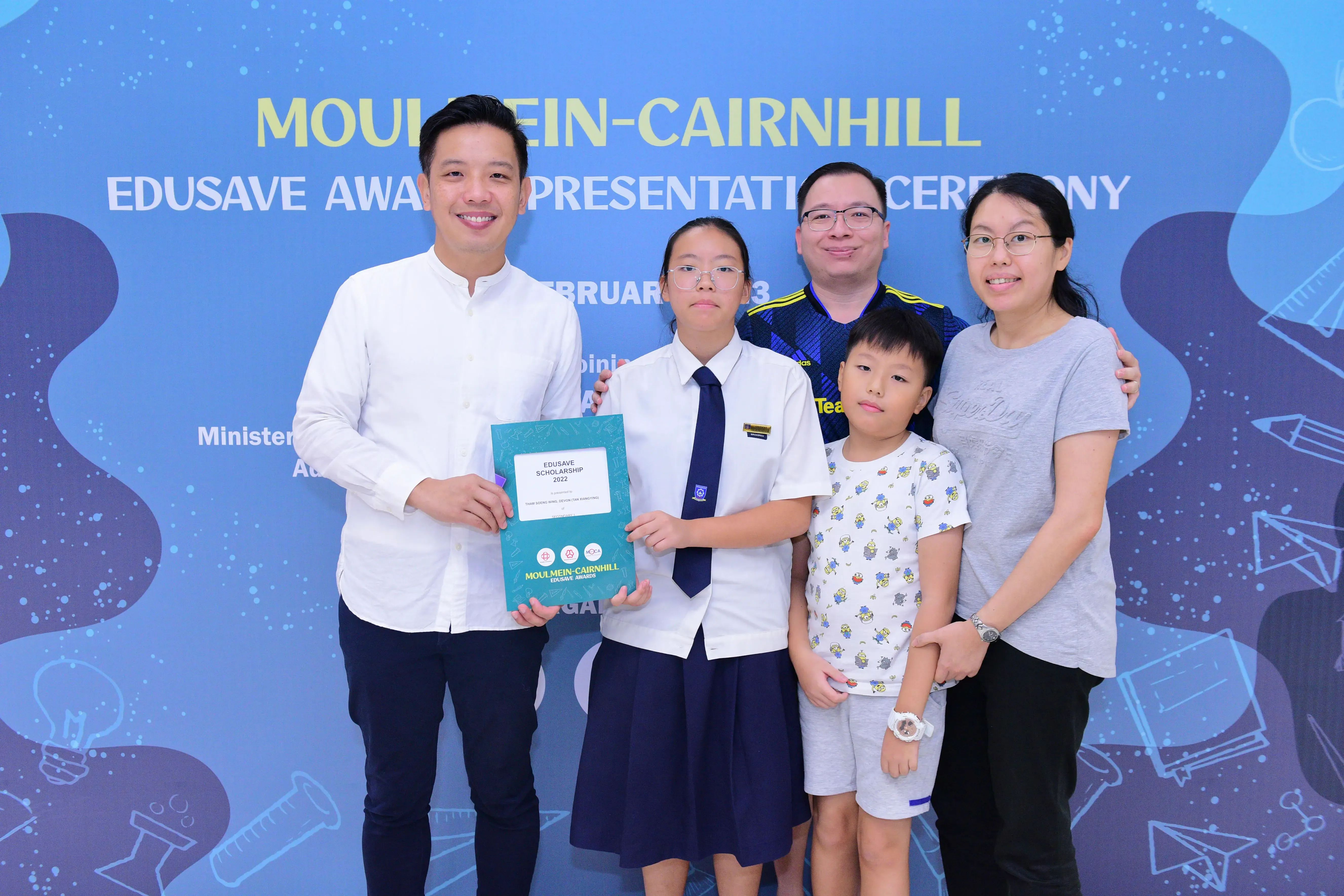 Five people pose at the Moulmein-Cairnhill Edusave Award Presentation Ceremony, with a student holding a scholarship certificate.