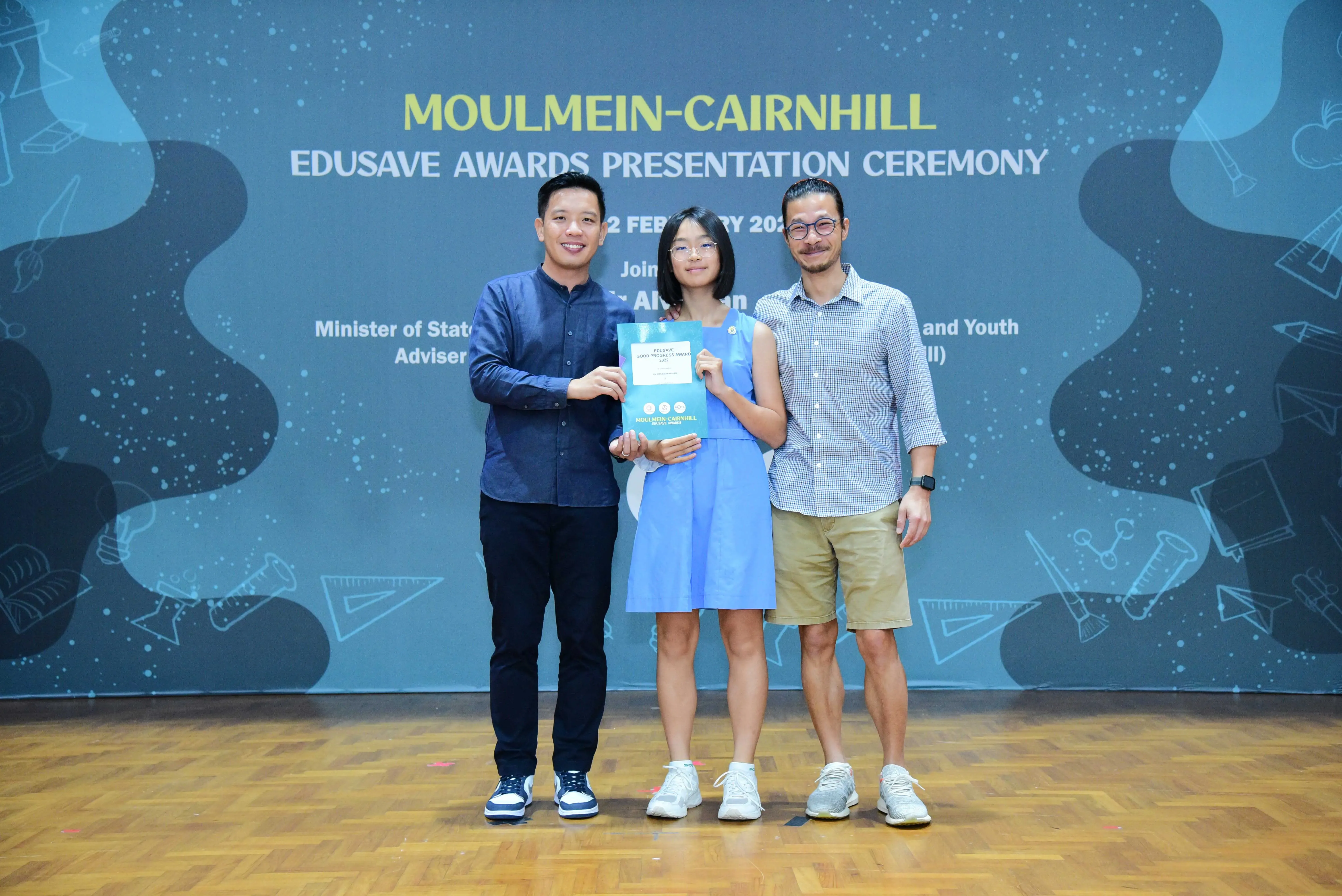 Three people pose holding a certificate at the Moulmein-Cairnhill Edusave Awards Presentation Ceremony backdrop.