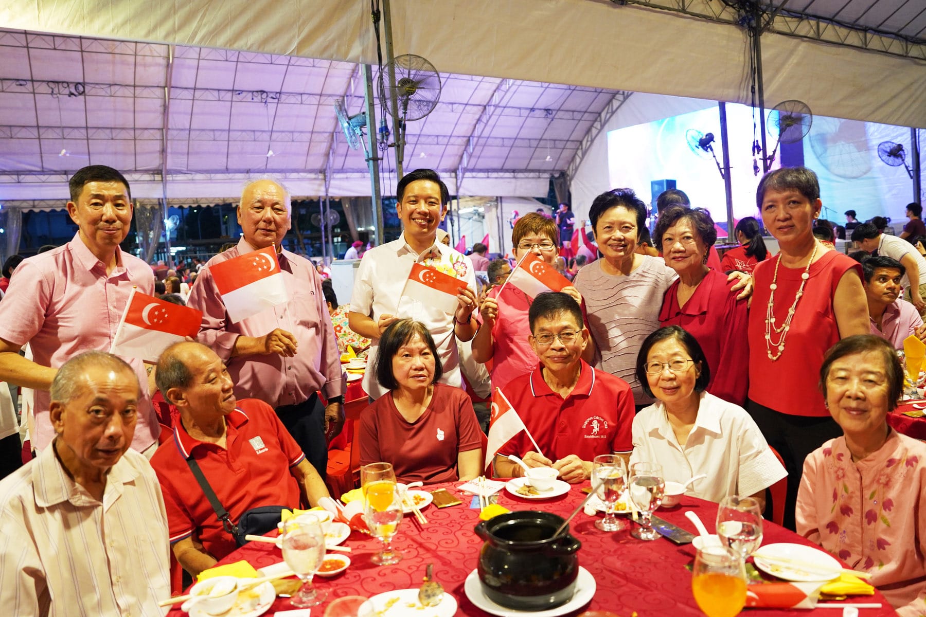 Group of people in red and white clothing holding Singapore flags at an outdoor dinner event under a tent.