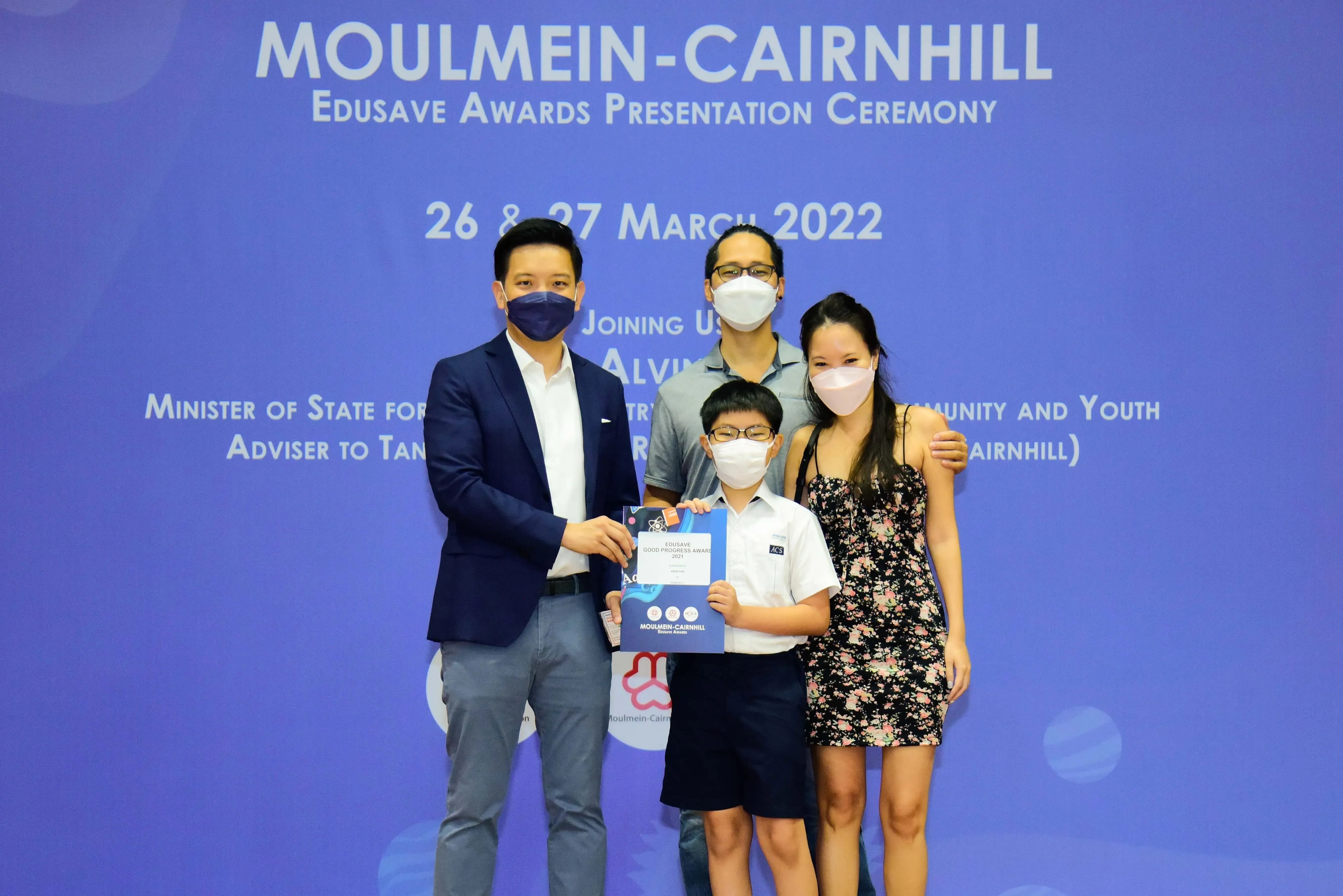 Four masked people pose at the Moulmein-Cairnhill Edusave Awards Presentation Ceremony, with a boy holding an award certificate.