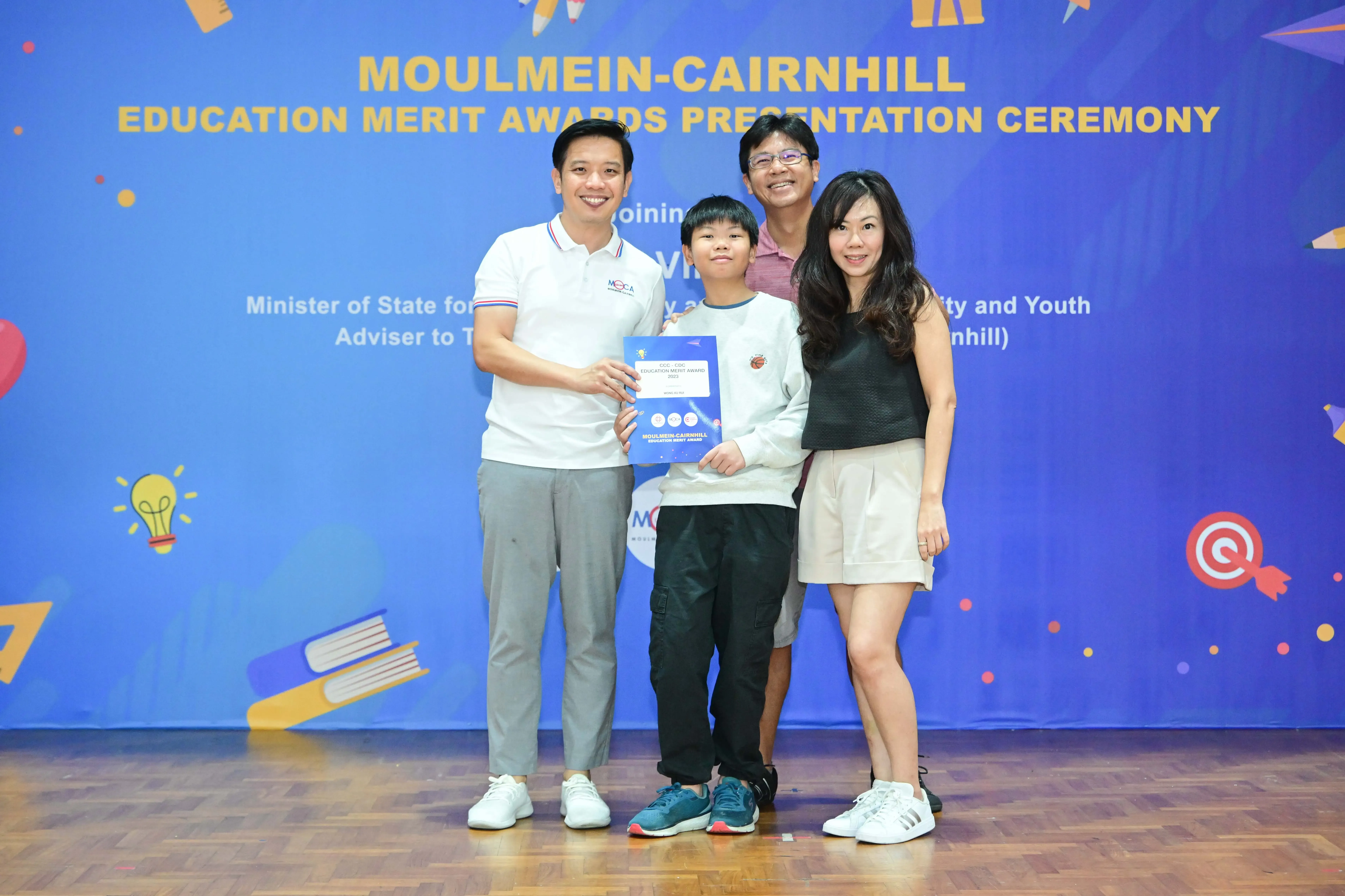 Four people pose with a certificate at the Moulmein-Cairnhill Education Merit Awards Presentation Ceremony banner.