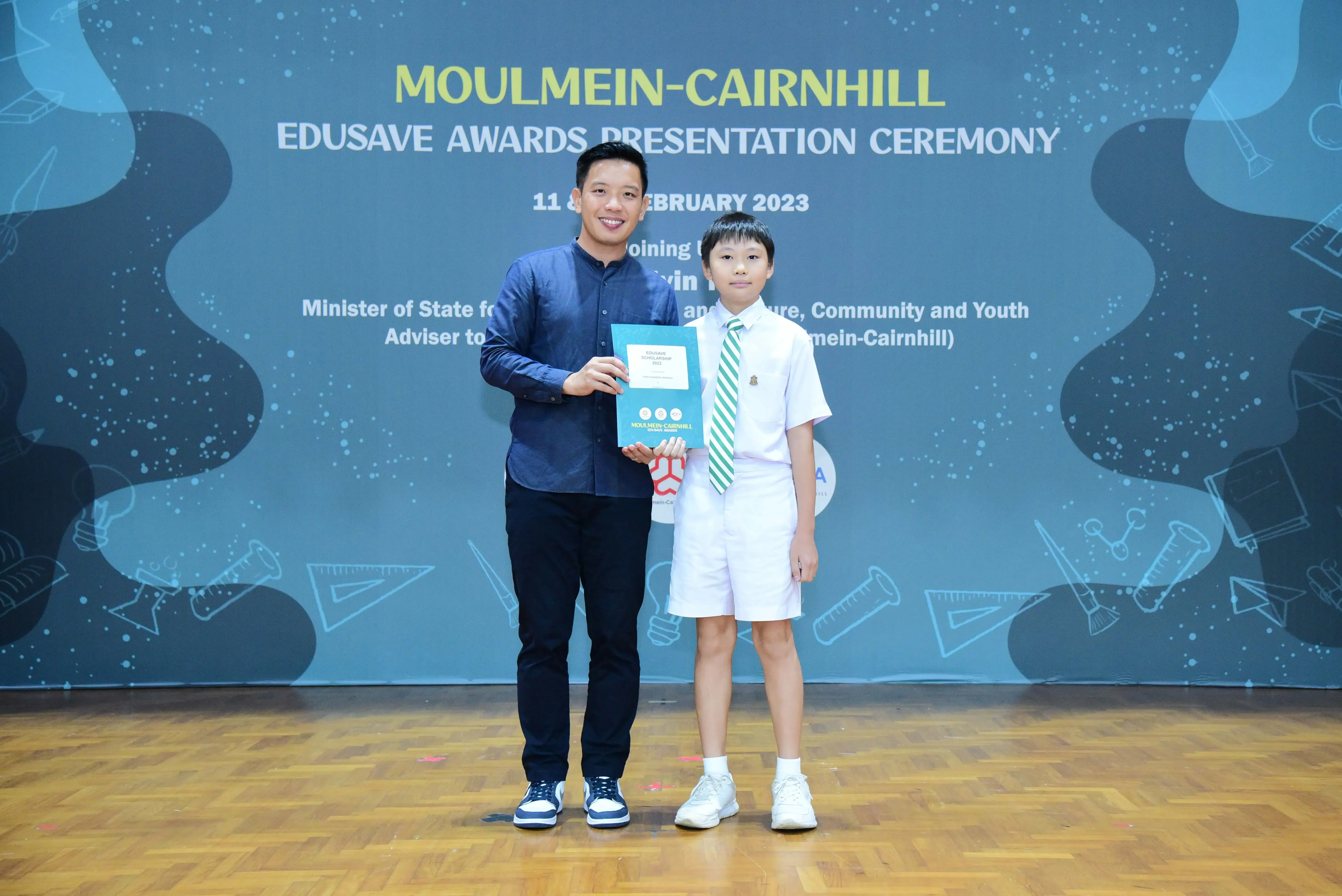 Man in blue shirt and young boy in school uniform pose holding an award at Moulmein-Cairnhill Edusave ceremony 2023.