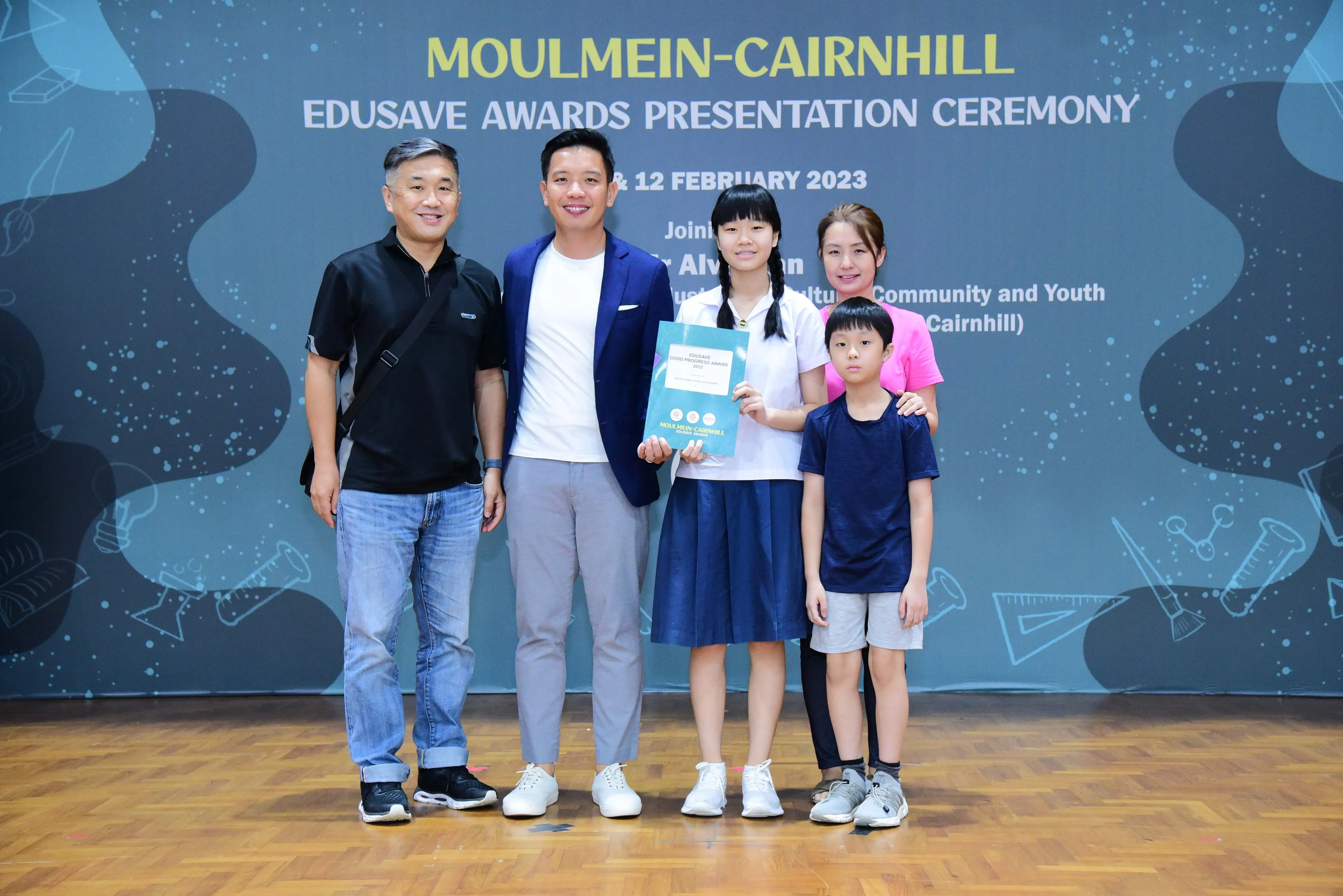 Five people pose in front of a Moulmein-Cairnhill Edusave Awards Presentation Ceremony banner, with a girl holding a certificate.