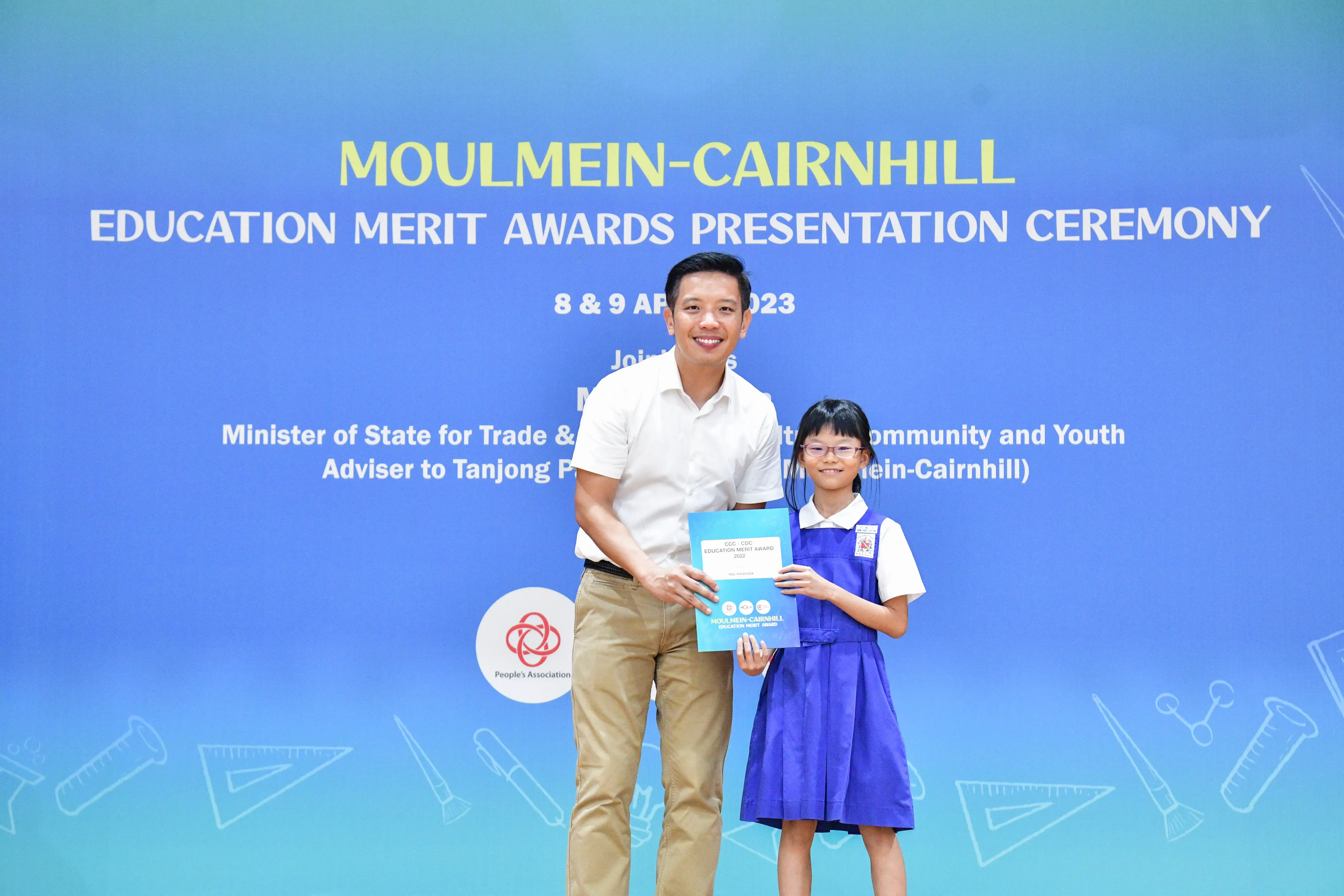 A man and a young girl hold an award certificate in front of a Moulmein-Cairnhill Education Merit Awards banner.
