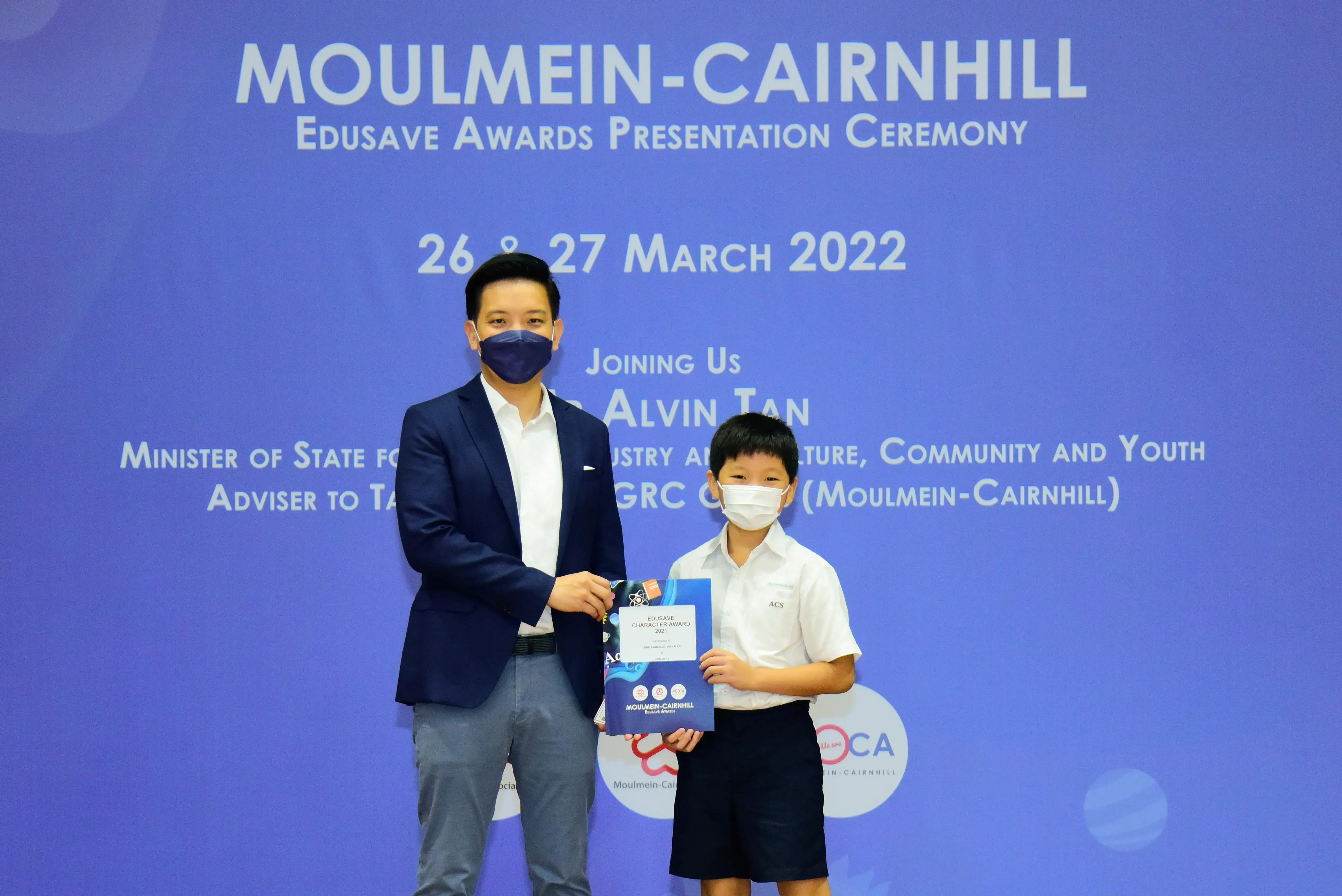 An adult and a young student in masks hold an award certificate at the Moulmein-Cairnhill Edusave Awards Ceremony.